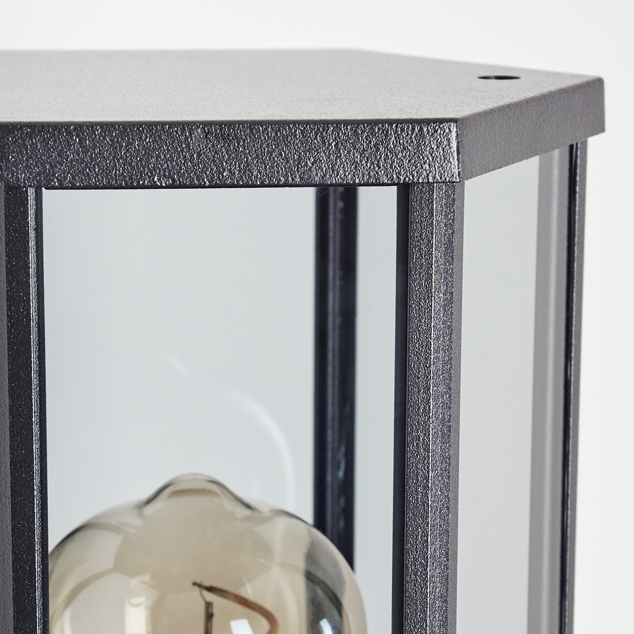 Nahaufnahme einer schwarzen, sechseckigen Lampe mit Glasscheiben. Im Inneren ist eine Glühbirne zu sehen. Der Hintergrund ist ein schlichtes Weiß.