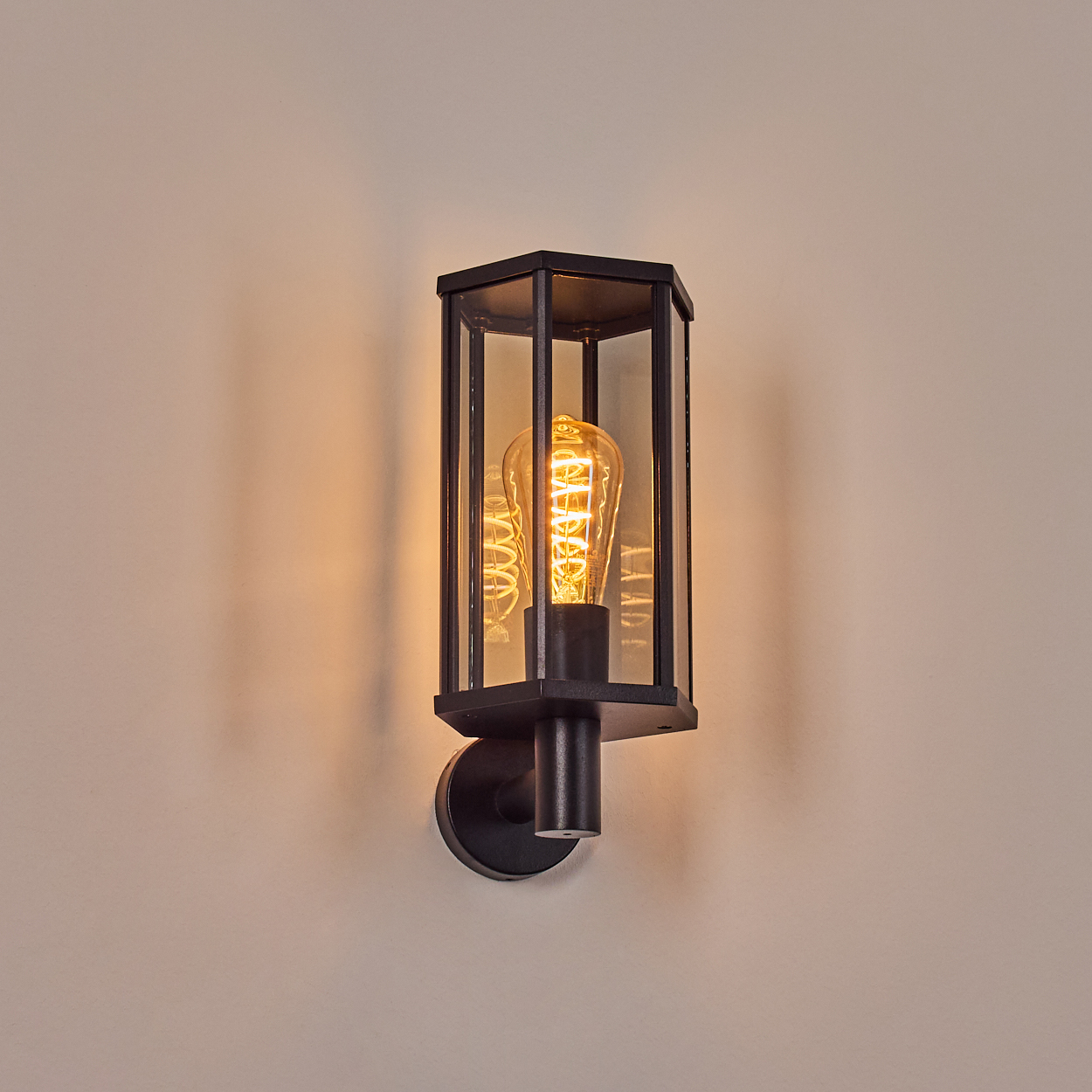 Schwarze Wandlampe mit Glaseinfassung und einer leuchtenden, warm getönten Glühbirne. Sie ist an einer hellbeigen Wand montiert.