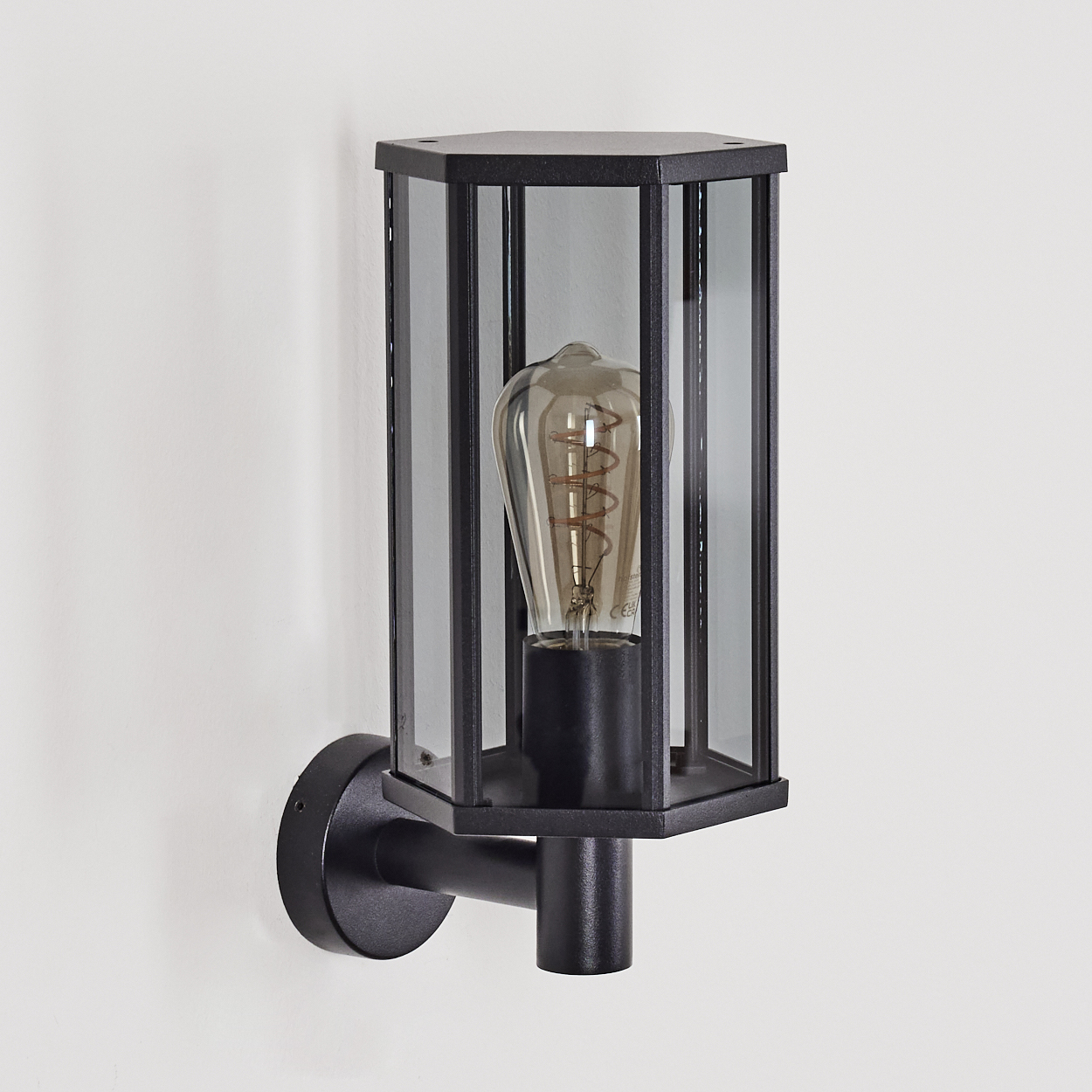 Schwarze Außenwandleuchte. Sechseckiges Glasgehäuse mit Glühbirne. An weißer Wand mit schwarzem Arm montiert.