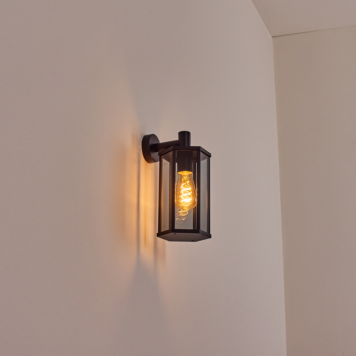 Schwarze Außenwandleuchte mit leuchtender Glühbirne, an einer beigen Wand montiert. Eine Ecke einer weißen Wand im Hintergrund.