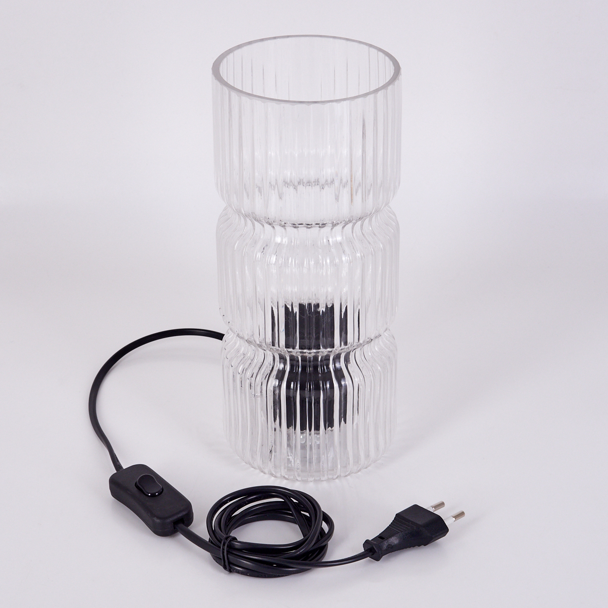 Eine klare, geriffelte Glaslampe mit einem schwarzen Netzkabel und Schalter. Die Lampe steht auf einer weißen Oberfläche.