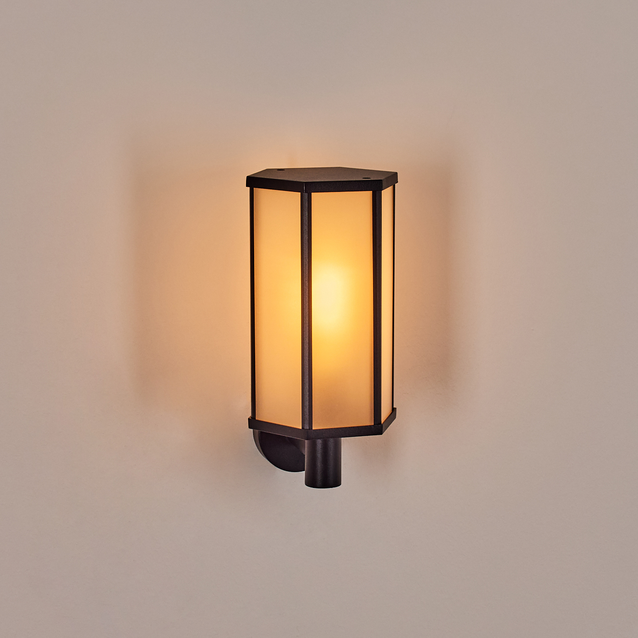 Eine Wandlampe. Sie hat einen schwarzen sechseckigen Rahmen mit Milchglasscheiben. Das Licht im Inneren ist warm und golden.