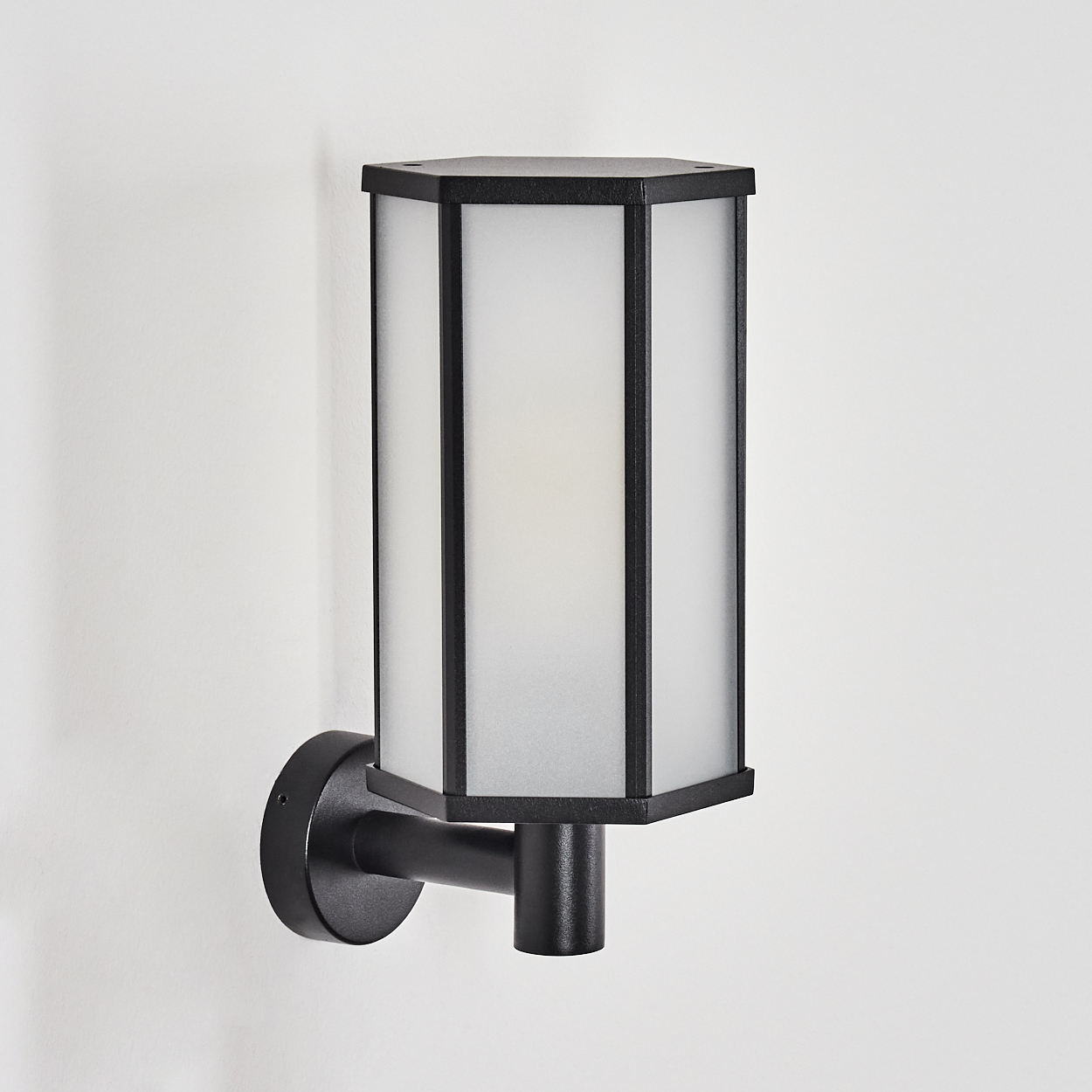 Eine schwarze Außenwandleuchte mit einem Schirm aus Milchglas, montiert an einer weißen Wand. Die Lampe hat ein geometrisches Design.