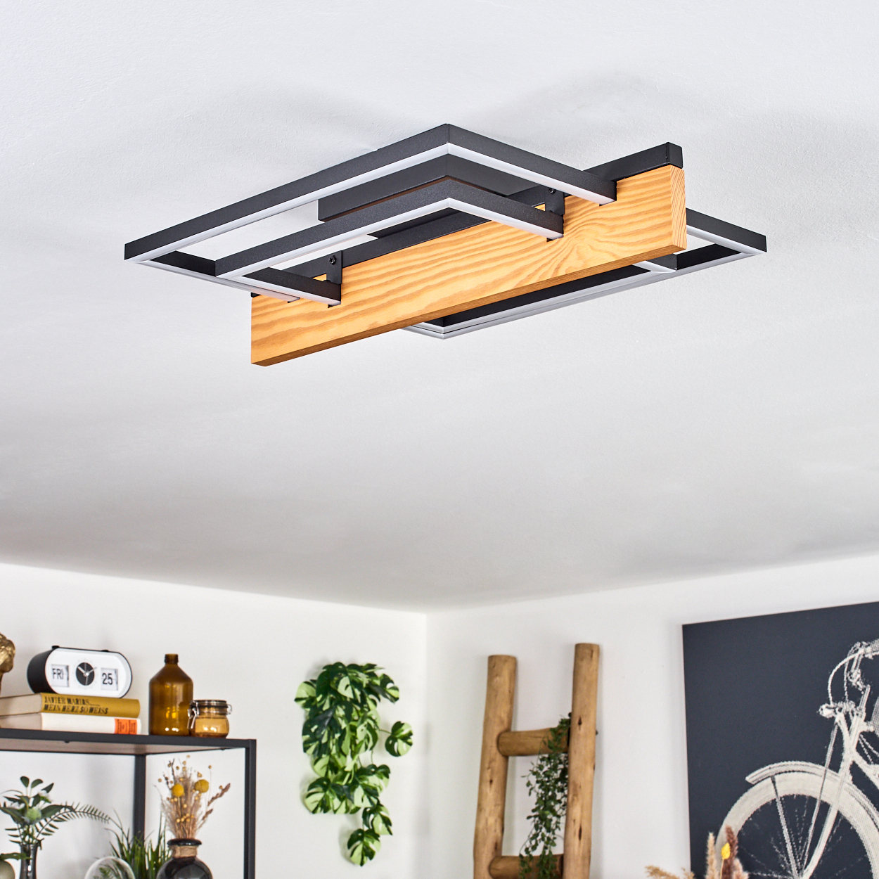Moderne Deckenlampe mit Holzbalken und schwarzen Rahmen. Der Hintergrund zeigt einen Raum mit Deko und einem Fahrradkunstwerk.
