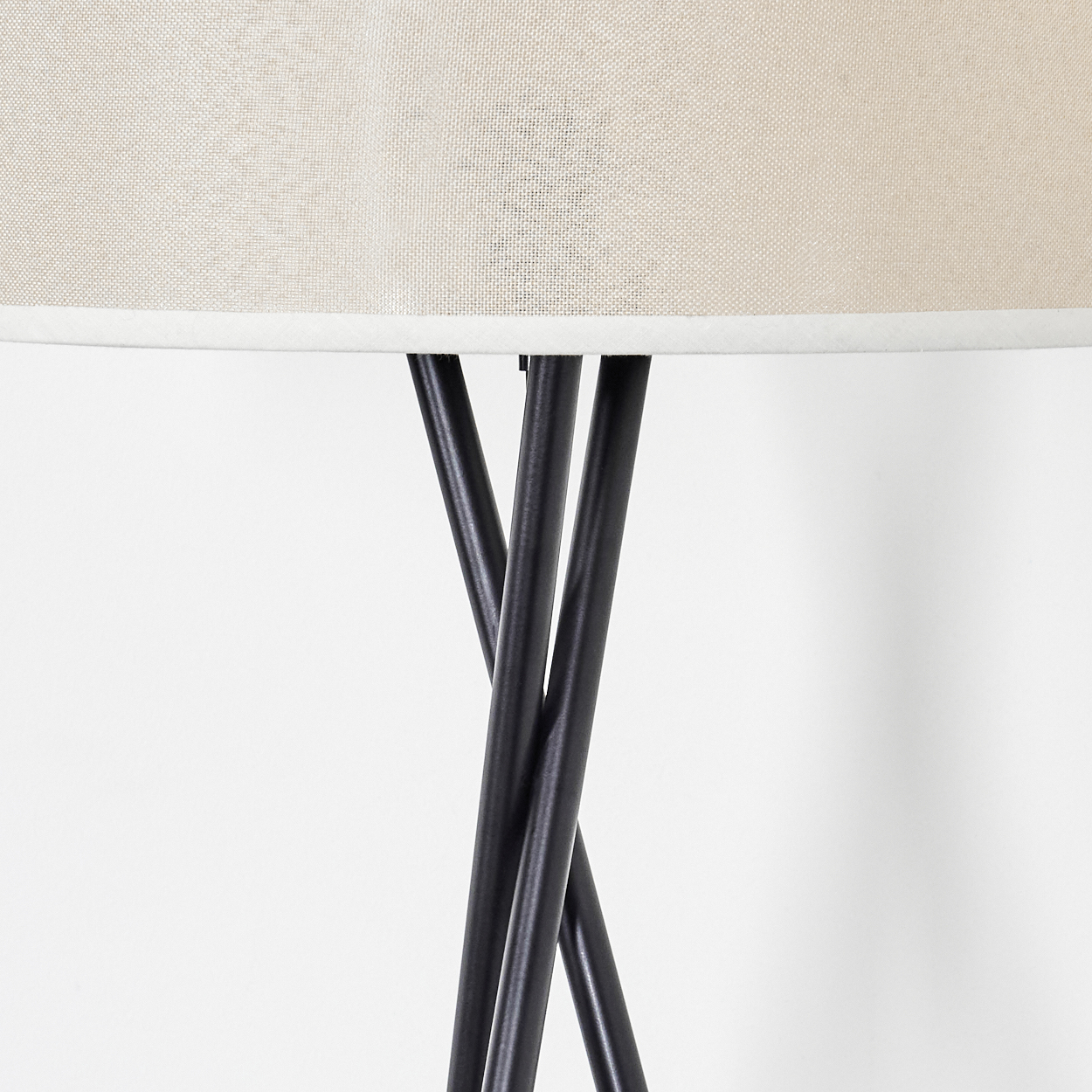 Nahaufnahme einer Lampe. Sie hat einen beigefarbenen Lampenschirm mit weißem Rand. Die Beine sind schwarz und kreuzen sich. Weißer Hintergrund.