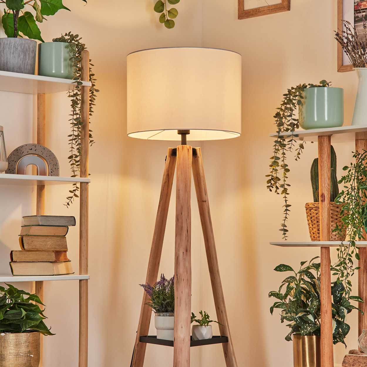 Eine hölzerne Stehlampe mit weißem Lampenschirm steht in einem Raum mit Regalen und Pflanzen. Bücher und Dekoration stehen auf den Regalen.