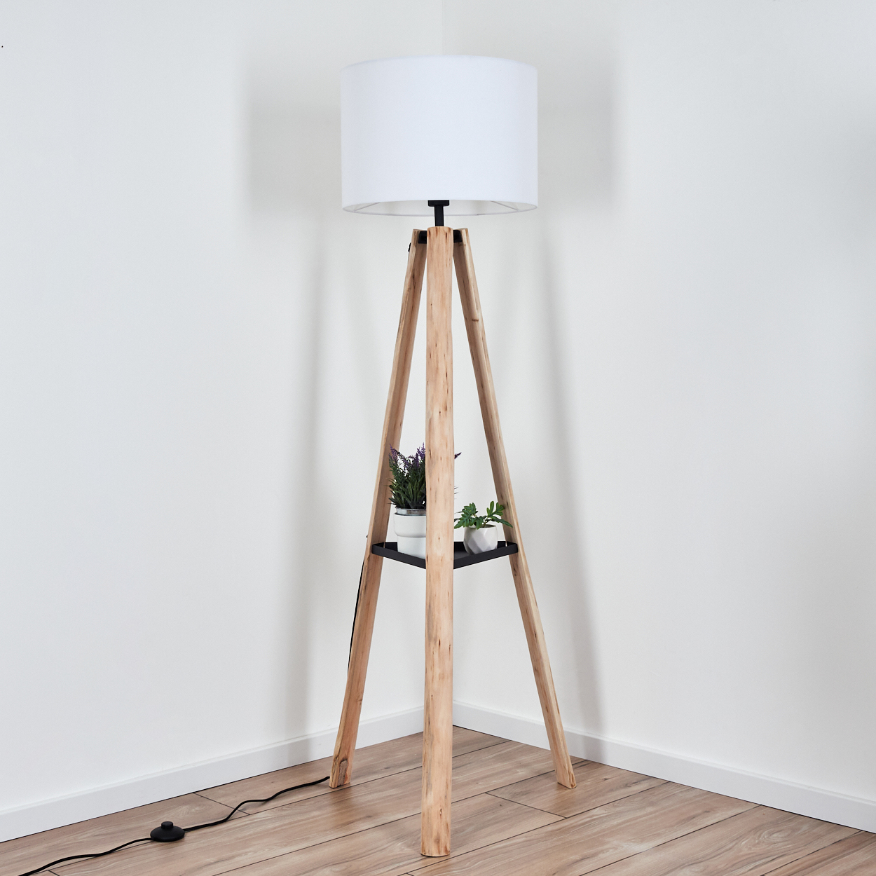 Eine hölzerne Stativ-Stehlampe mit weißem Lampenschirm. Ein schwarzes Regal hält Topfpflanzen. Die Lampe steht in einer Ecke.