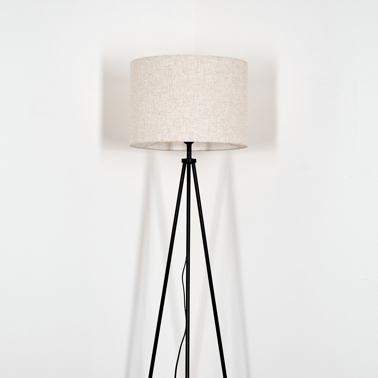 Eine Stehlampe mit beigefarbenem Lampenschirm und schwarzem Stativfuß steht vor einer weißen Wand. Die Lampe wirft einen Schatten.