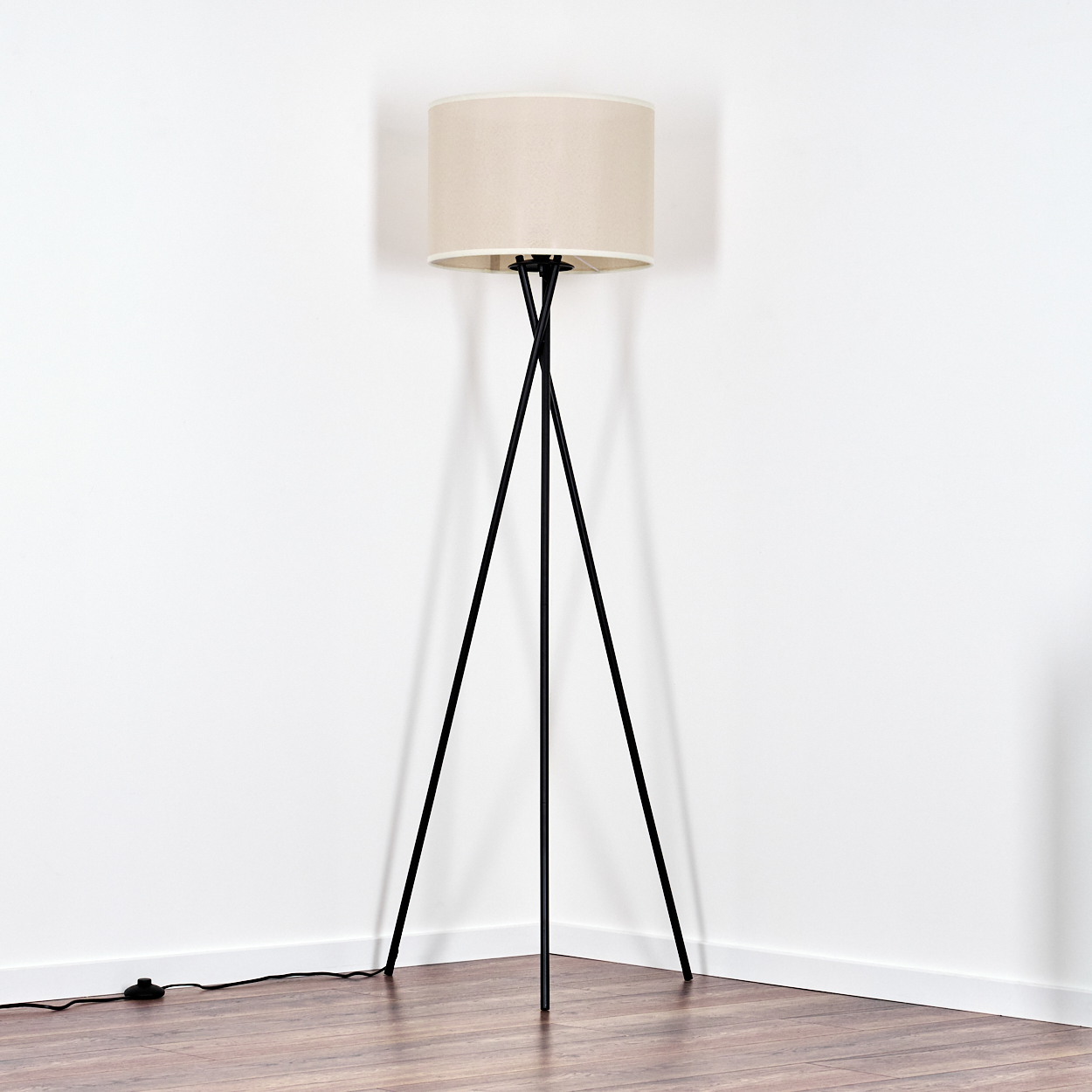 Eine Stehlampe mit beigem Lampenschirm und schwarzen Dreibeinbeinen steht in der Ecke eines Raumes mit weißen Wänden und Holzboden.