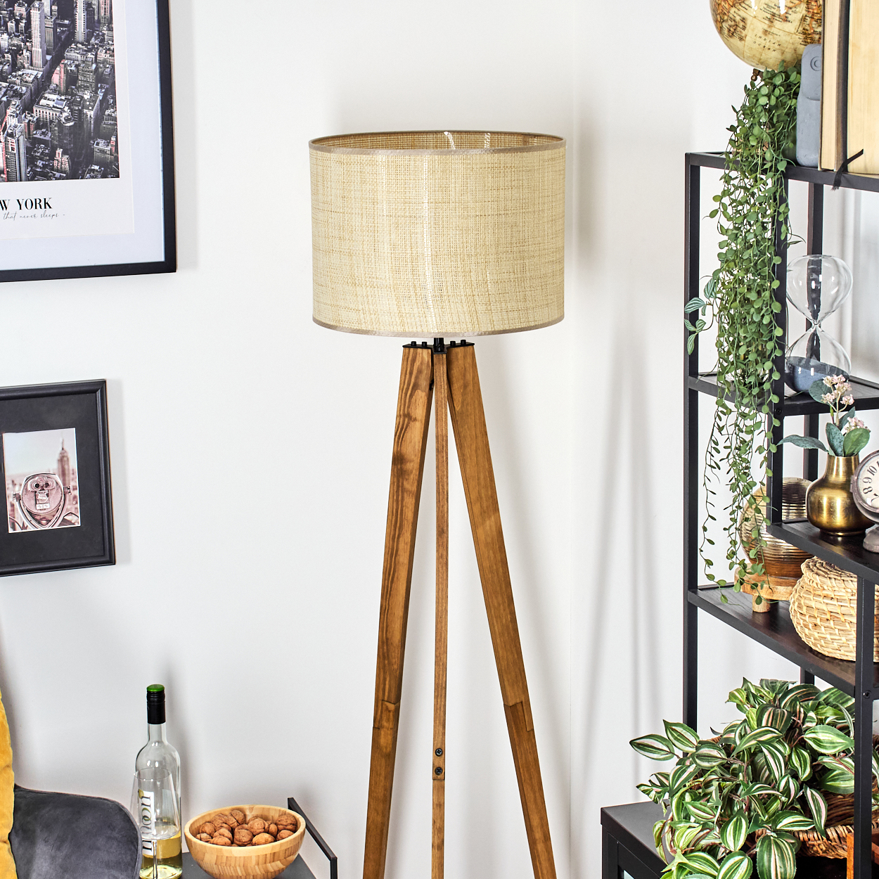 Eine hölzerne Stehlampe mit beigem Schirm steht in einem Raum. Regale mit Pflanzen und Dekoration sind rechts. Bilder und eine Schale sind links.