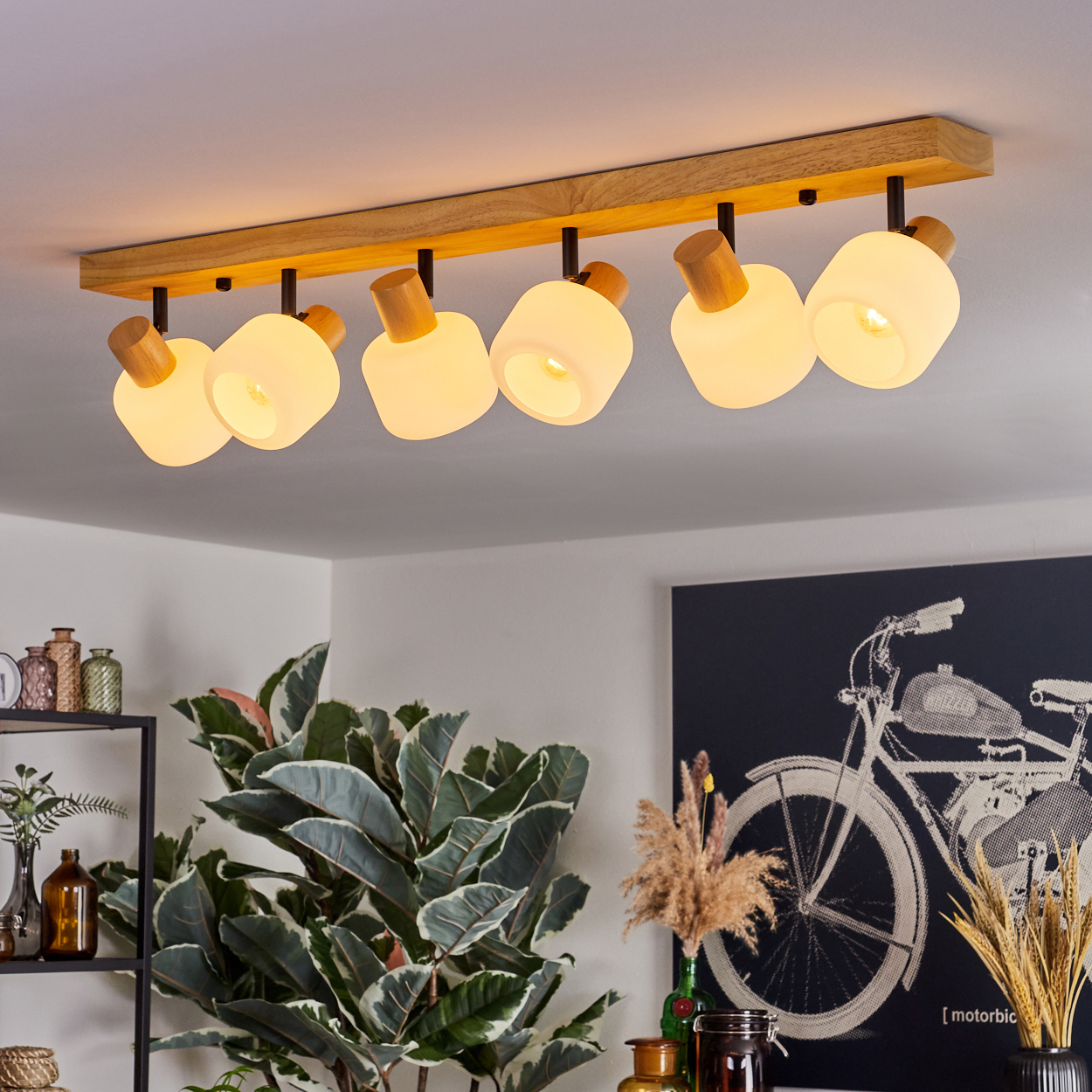 Eine Deckenleuchte mit sechs Lampen. Holzbalken, weiße Lampenschirme. Eine Pflanze und ein Fahrrad-Kunstwerk im Hintergrund.
