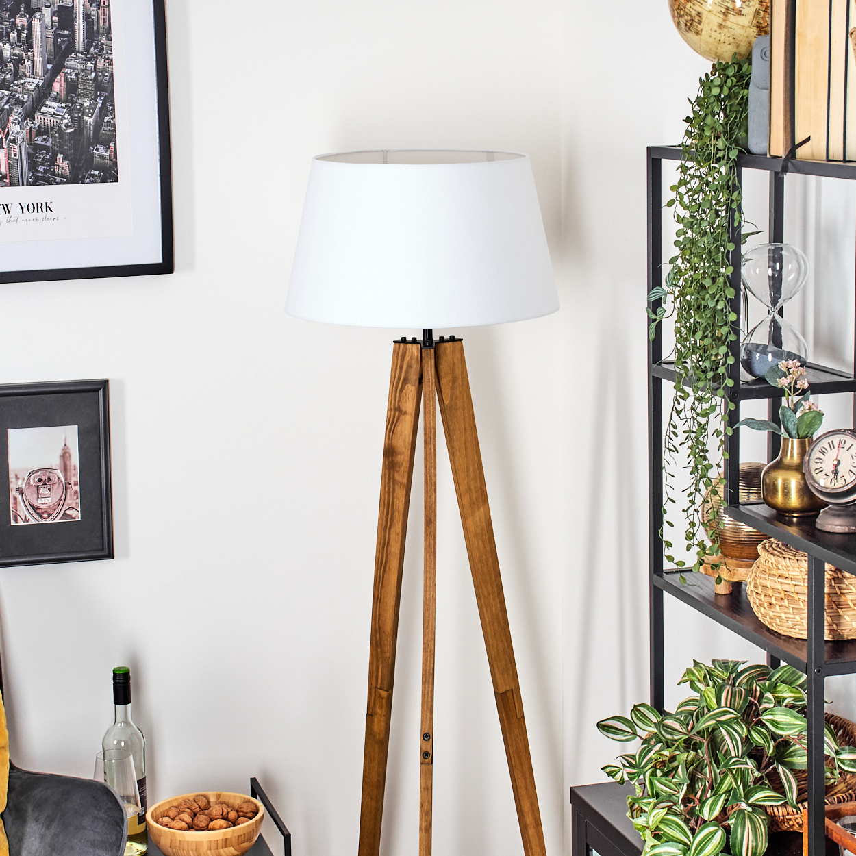 Eine hölzerne Stativlampe mit weißem Schirm steht neben einem schwarzen Regal mit Dekoration. An der Wand hängt ein gerahmtes New Yorker Stadtbild.