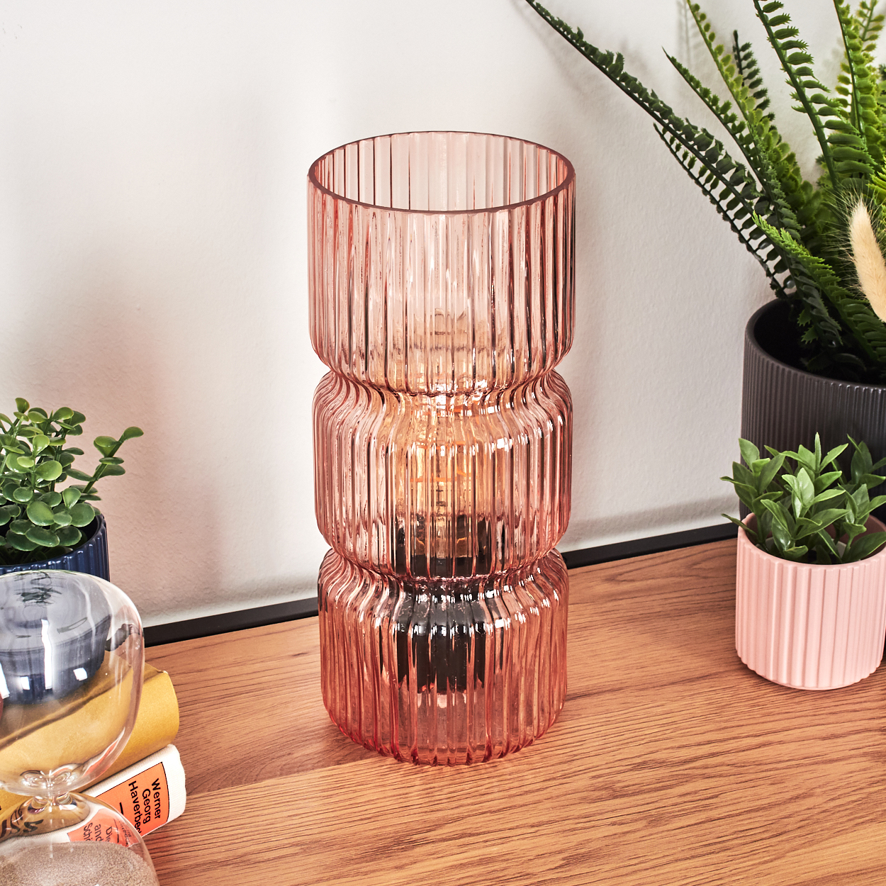 Eine rosa Glaslampe steht auf einer Holzoberfläche. Pflanzen und eine Sanduhr sind im Hintergrund. Die Lampe ist beleuchtet.