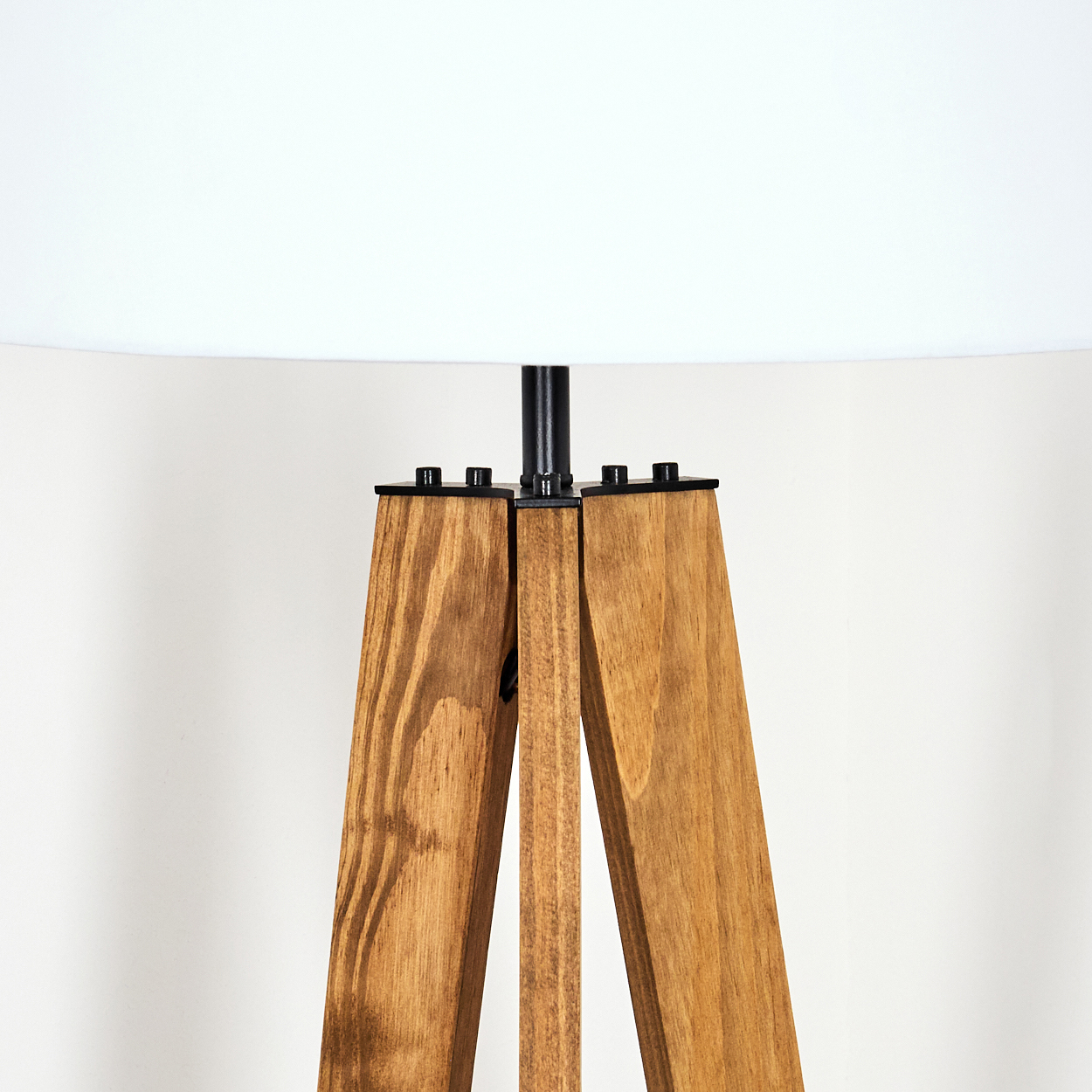 Gezeigt wird eine Stehlampe mit Holzstativ. Die Lampe hat einen weißen Lampenschirm und eine schwarze Metallstange. Die Beine sind aus braunem Holz.