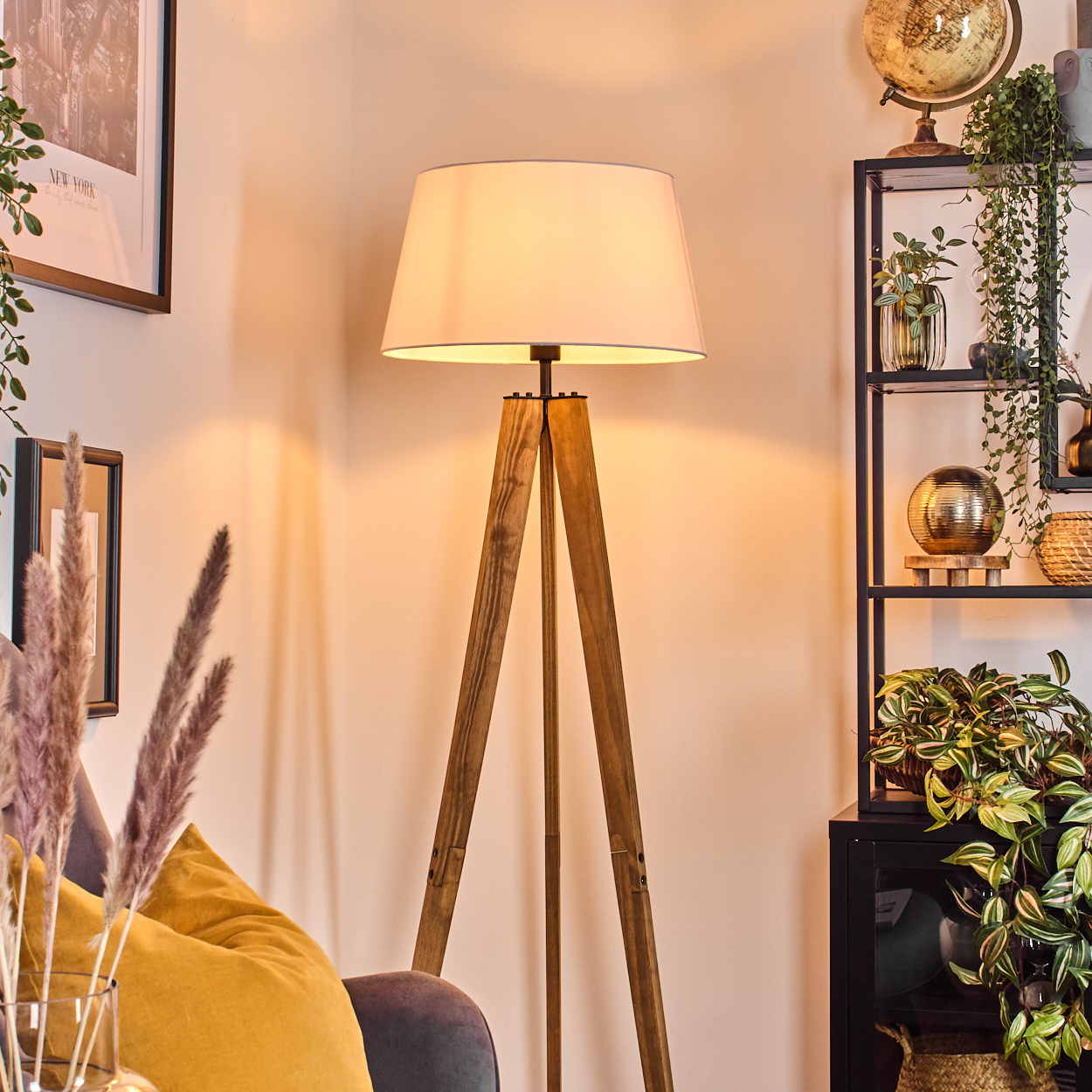 Eine Holz-Stativlampe mit Lampenschirm, ein gelbes Kissen, Pflanzen, ein Regal und ein Globus. Die Szene hat warmes Licht.