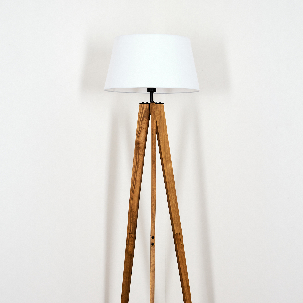 Eine Stehlampe aus Holz mit weißem Lampenschirm steht vor einer weißen Wand. Die Lampe hat ein modernes Design.