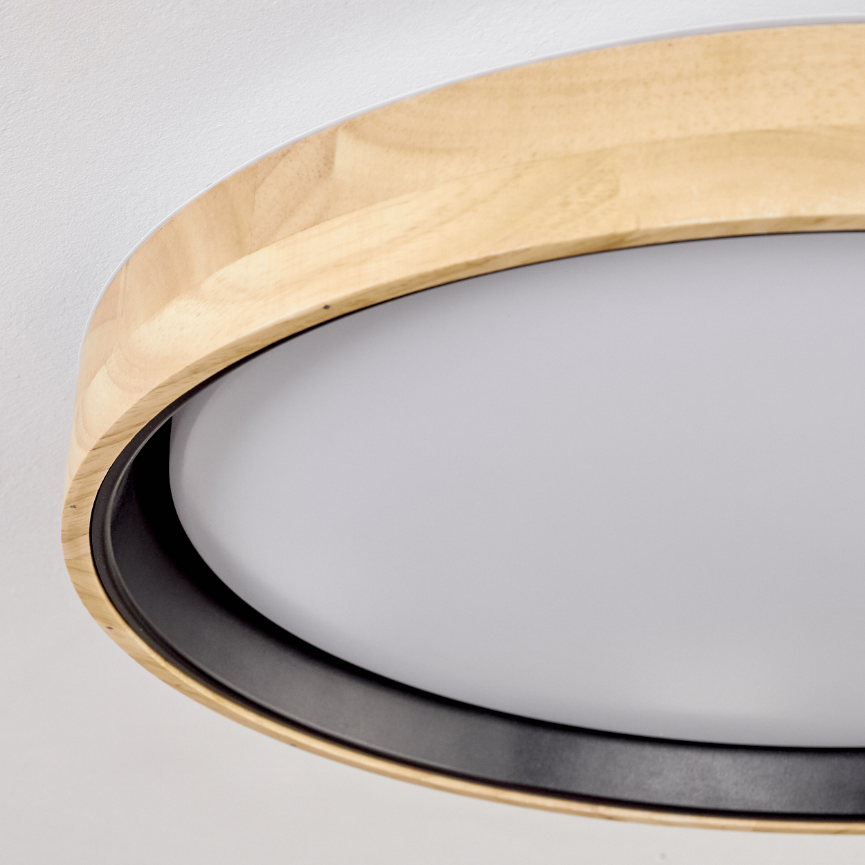 Nahaufnahme einer runden Deckenleuchte. Sie hat einen Holzrahmen, einen schwarzen Ring und einen weißen Lichtdiffusor. Der Hintergrund ist weiß.