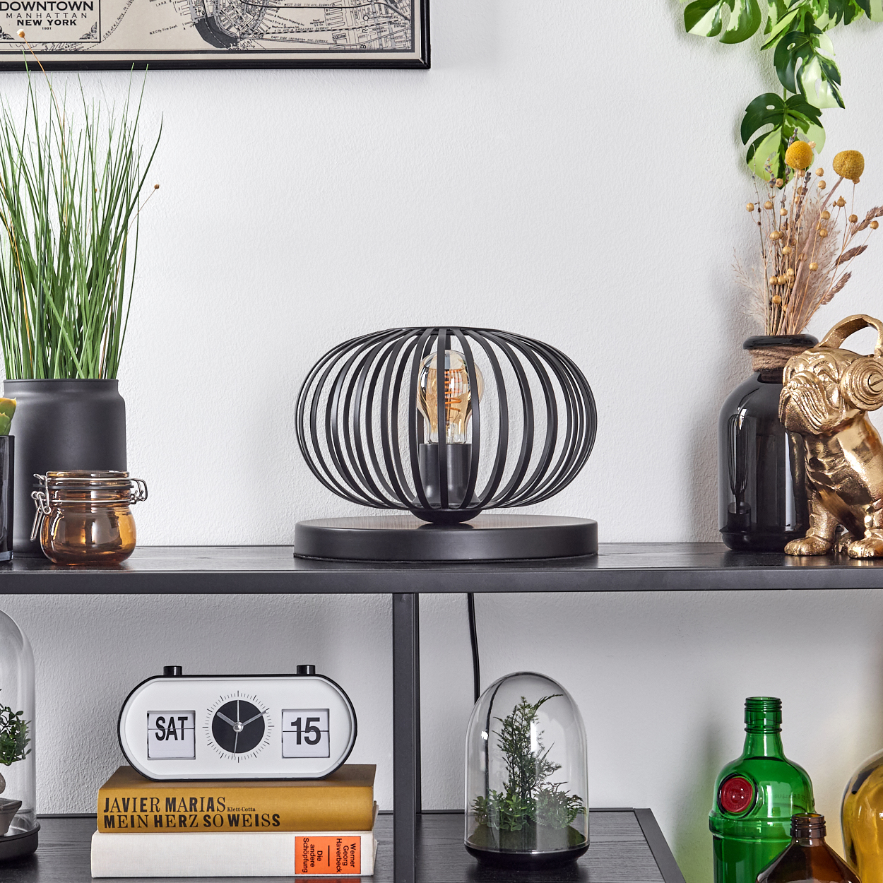 Eine schwarze Metalllampe mit einer Glühbirne steht auf einem Regal mit Dekorationen. Eine Uhr, ein Buch und andere Gegenstände sind ebenfalls auf dem Regal.