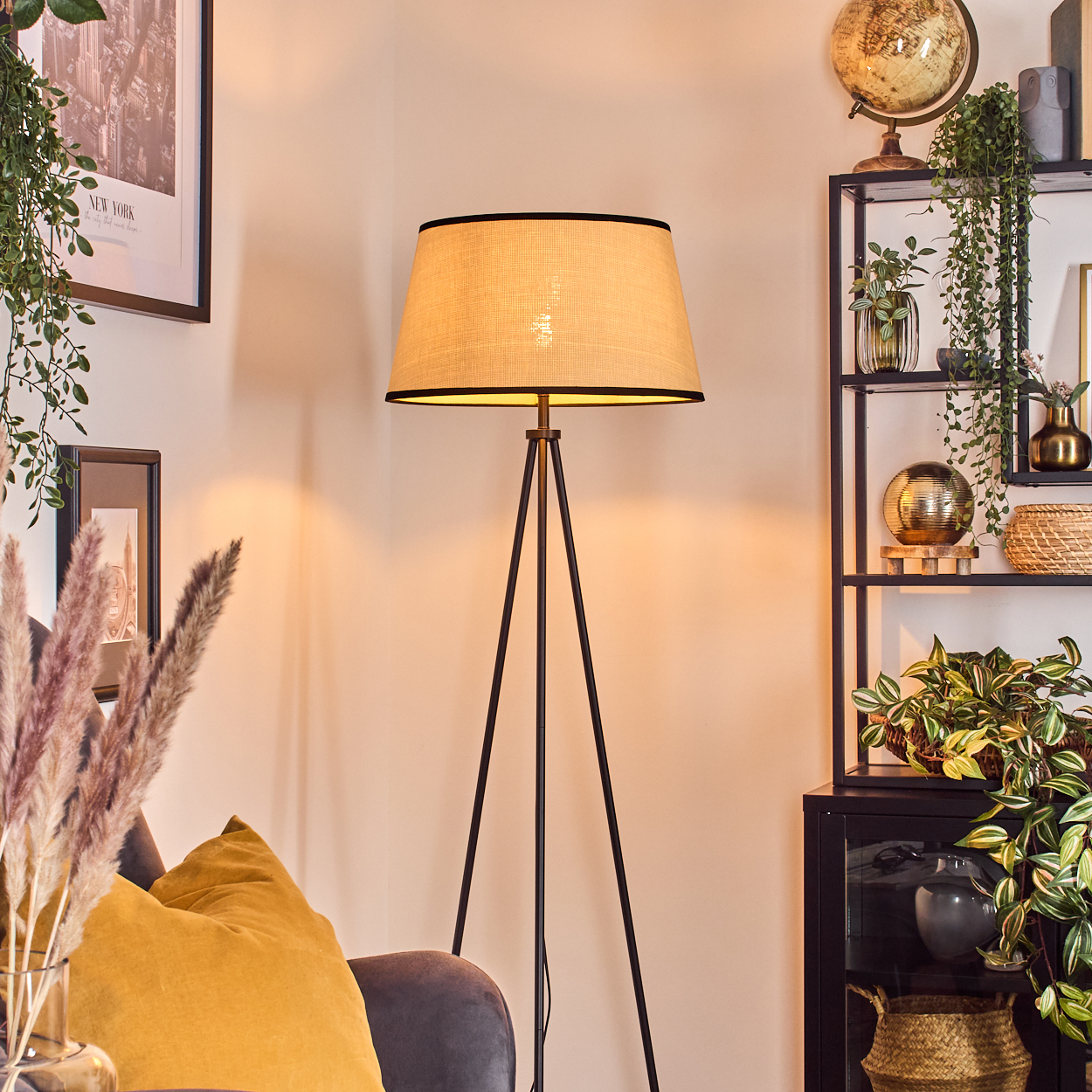 Eine beleuchtete Stehlampe steht neben einem Regal mit Pflanzen und Dekoration. Ein gelbes Kissen liegt auf einem grauen Stuhl.