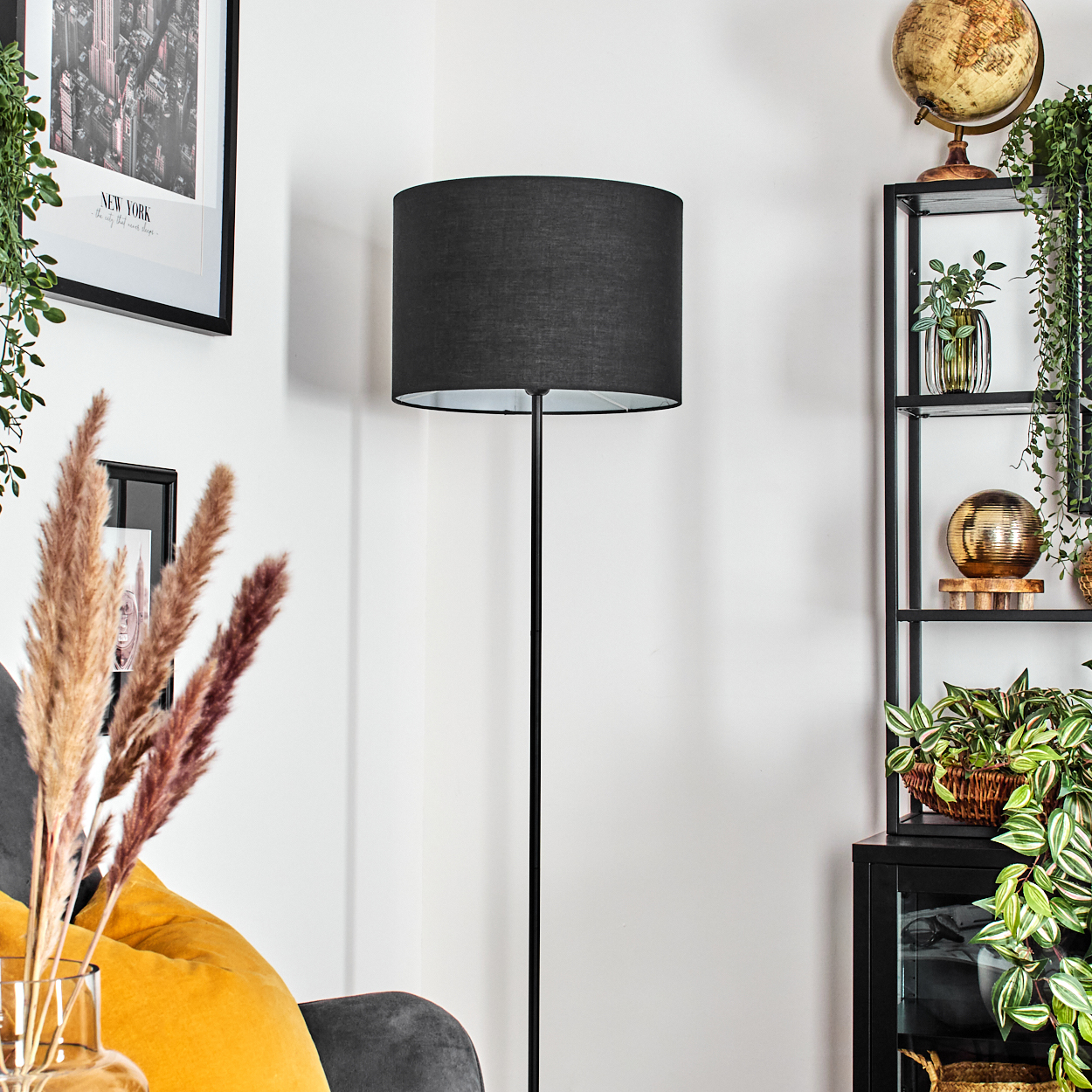 Eine schwarze Stehlampe steht in einem Raum. Rechts ein Regal mit Pflanzen. Links ein Bild und Pampasgras.