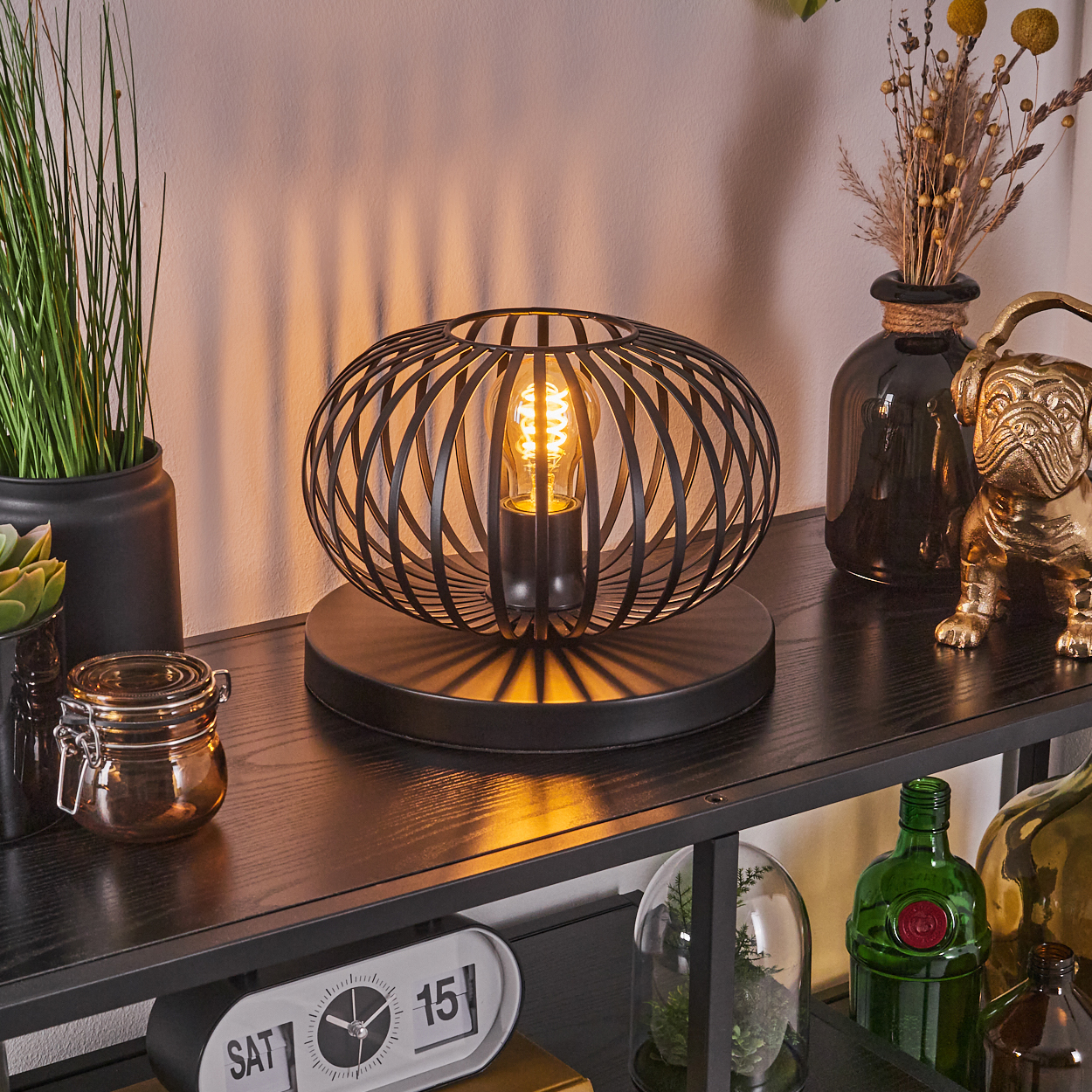 Eine schwarze Metalllampe mit leuchtender Glühbirne steht auf einem Regal mit Dekoration. Eine goldene Bulldoggenstatue und Vasen sind ebenfalls vorhanden.