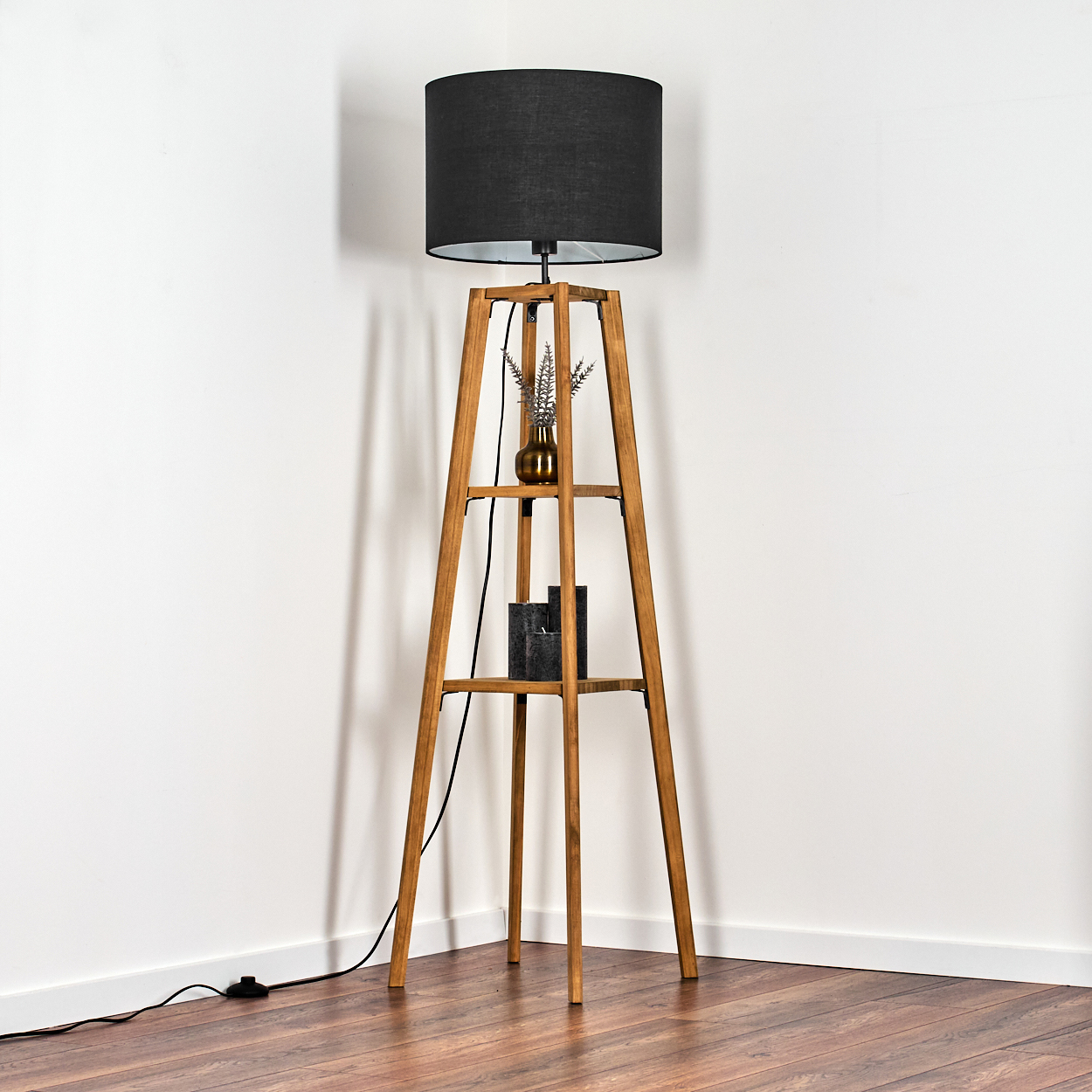 Stehlampe aus Holz mit Ablagen. Sie hat einen schwarzen Lampenschirm, eine kleine Vase und zwei schwarze Dekorationsgegenstände. Der Hintergrund ist eine weiße Wand.