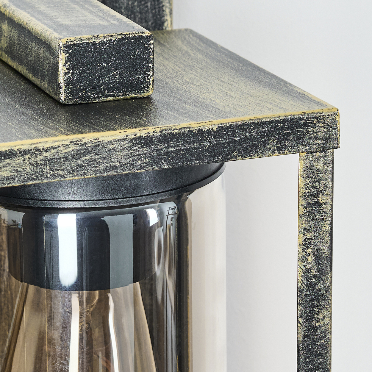 Nahaufnahme einer schwarz-goldenen Wandleuchte. Sie hat einen Glaszylinder und einen Holzrahmen. Der Hintergrund ist weiß.