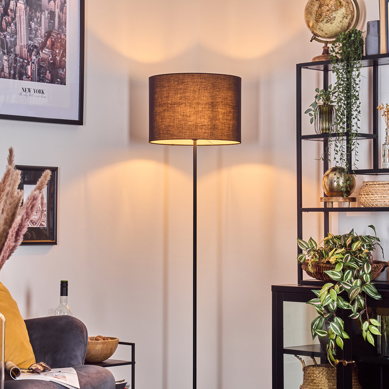 Ein Raum mit Stehlampe, Regalen mit Pflanzen und Globus, Sofa und New York Skyline-Poster.