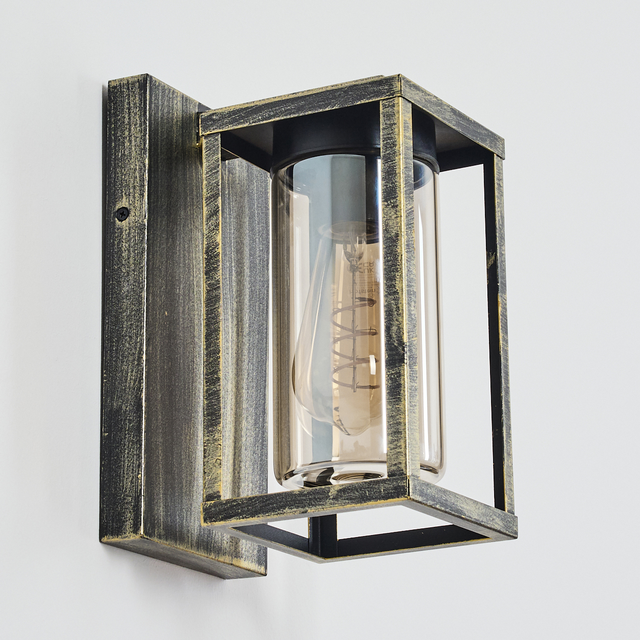 Wandleuchte mit Glaszylinder und Edison-Glühbirne, umgeben von einem schwarz-goldenen Metallrahmen, an einer weißen Wand.