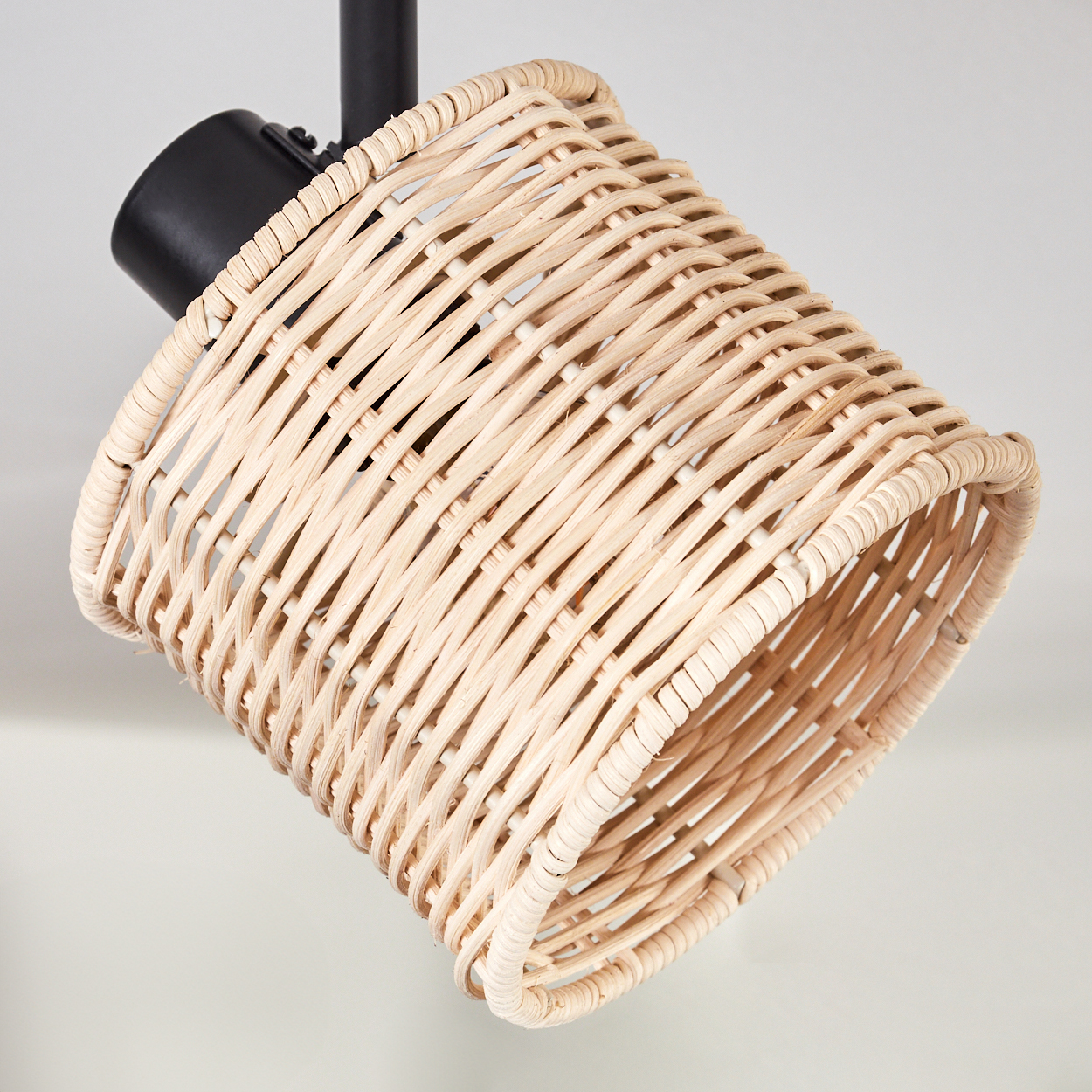 Ein Lampenschirm aus geflochtenem Rattan, zylindrische Form, hängt an einer schwarzen Halterung vor weißem Hintergrund.