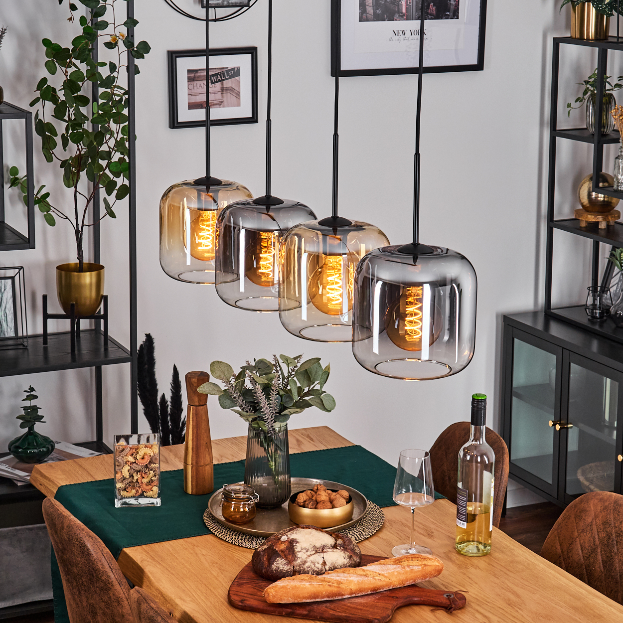 Esszimmer mit Hängelampen über einem Holztisch, gedeckt mit Essen, Wein und einem grünen Läufer. Schwarze gerahmte Kunst und Regale sind sichtbar.