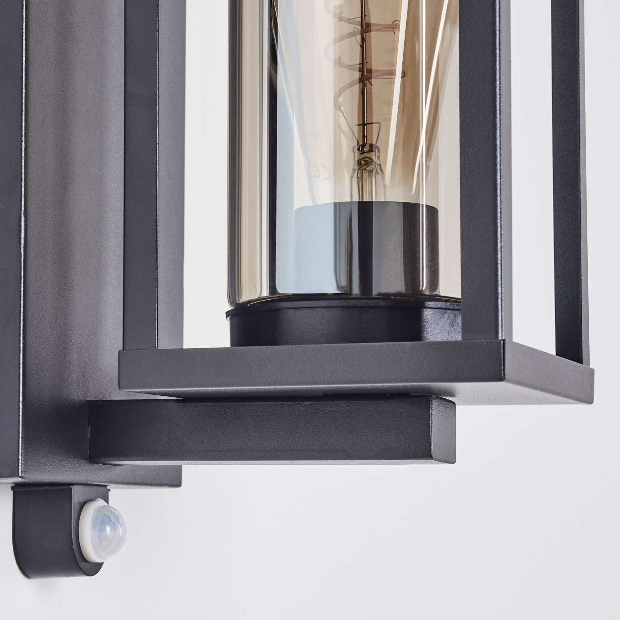 Nahaufnahme einer schwarzen Metall-Außenlampe mit Glühbirne. Auf der linken Seite ist ein Bewegungssensor an einer weißen Wand zu sehen.