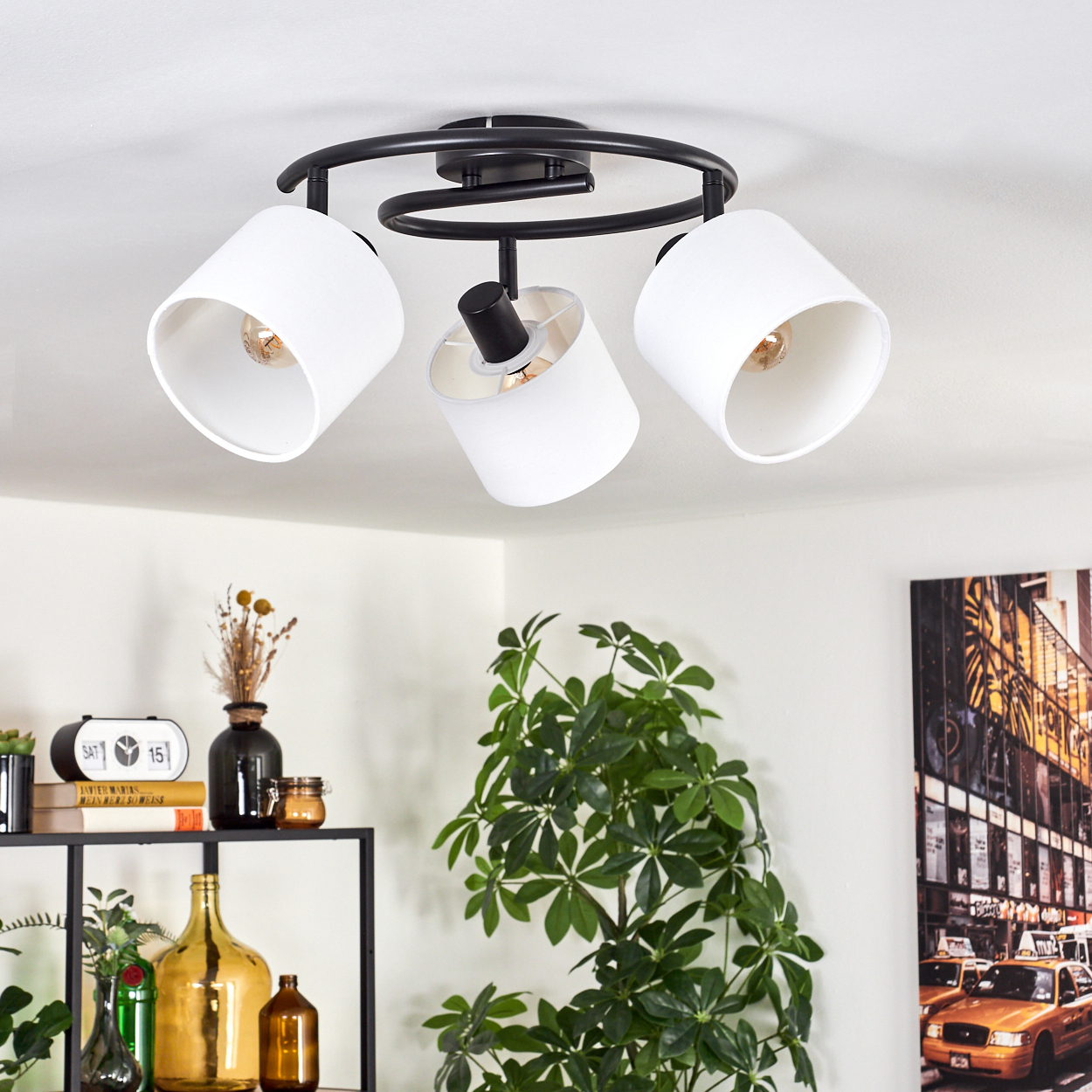 Deckenleuchte mit drei weißen Schirmen. Darunter ein Regal mit Deko, eine Pflanze und ein New York City-Foto an der Wand.