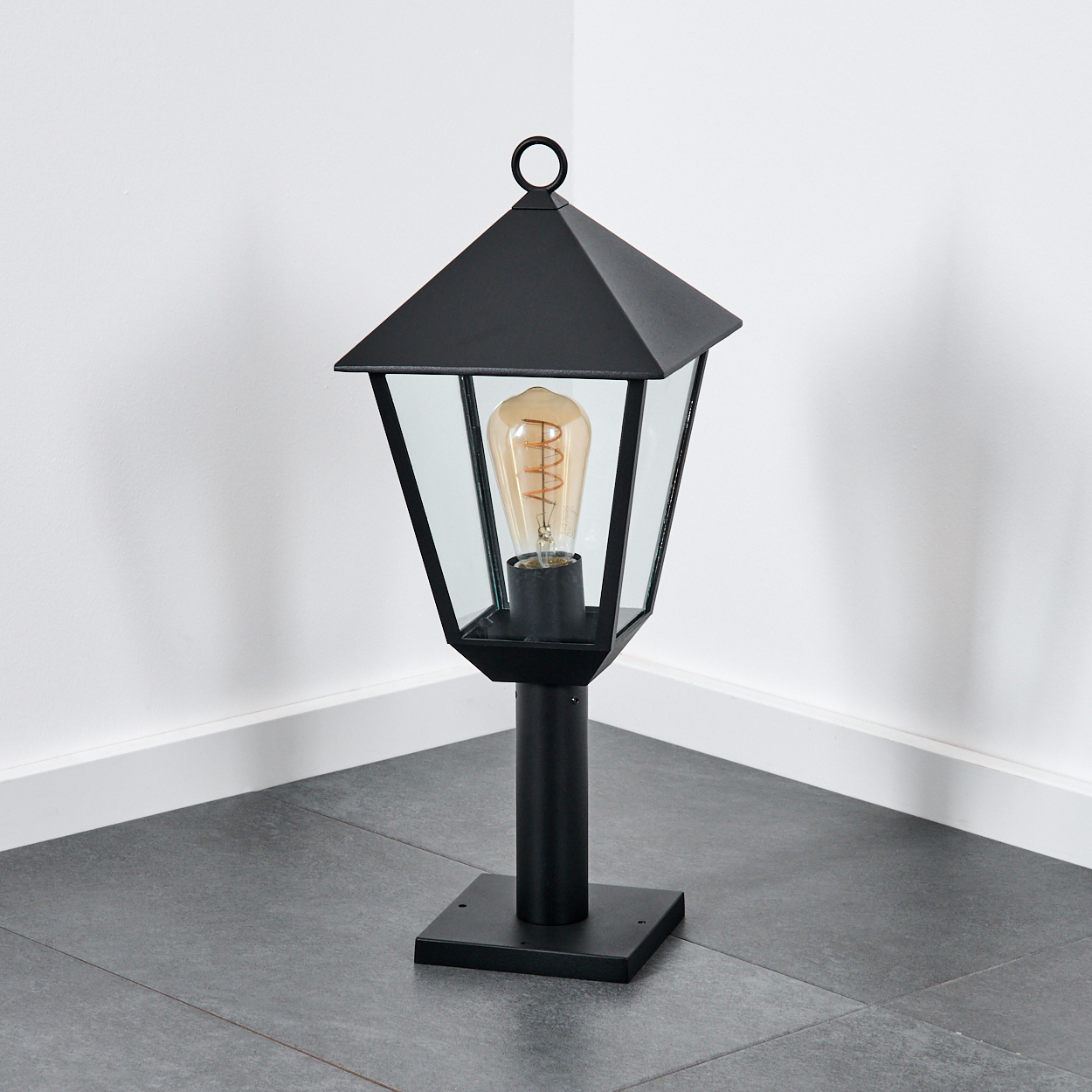 Schwarze Außenlampe mit Glasgehäuse. Eine brennende Glühbirne ist darin. Sie steht auf einem schwarzen Sockel. Weiße Wand und grauer Boden im Hintergrund.