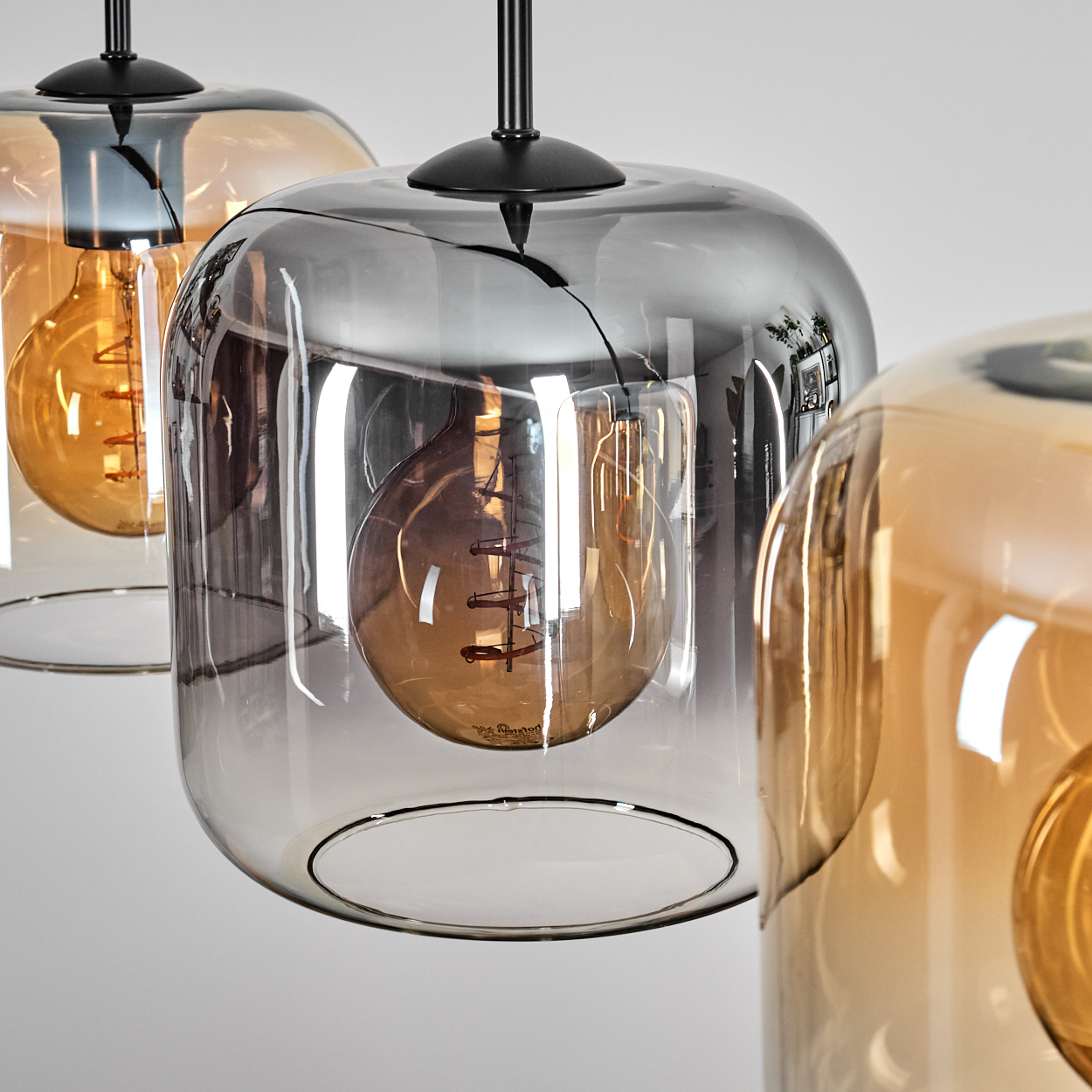 Drei moderne Pendelleuchten mit Glasschirmen und freiliegenden Glühbirnen. Die Schirme sind grau und bernsteinfarben, mit schwarzen Metallakzenten.