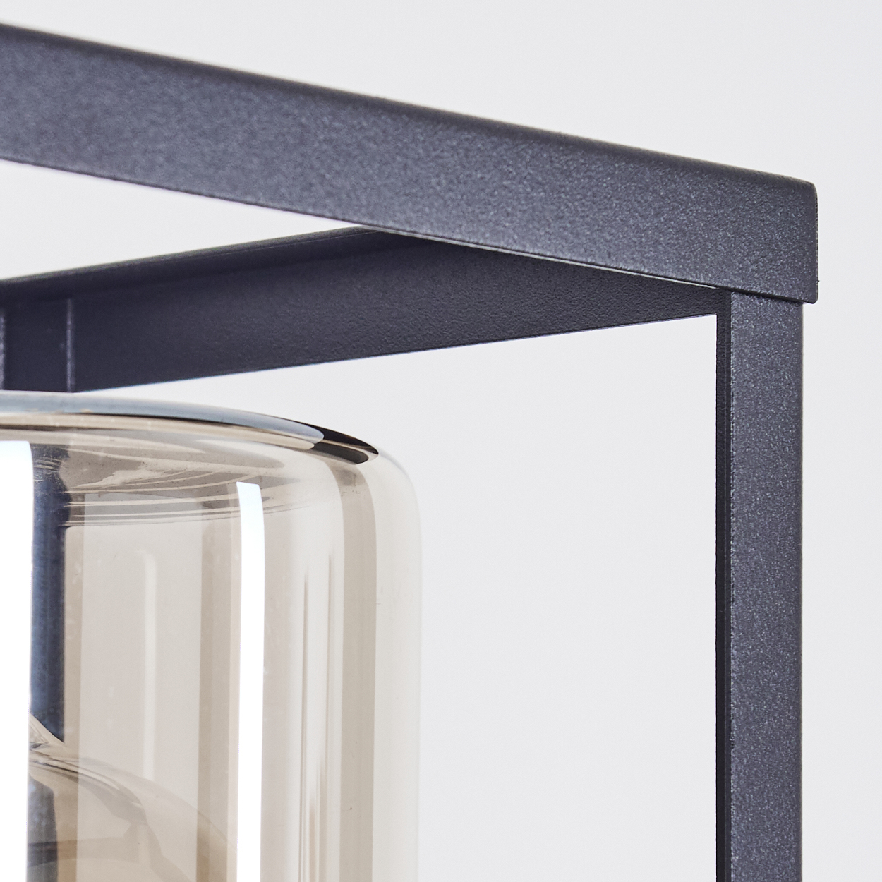 Nahaufnahme eines dunkelgrauen Metallrahmens mit einem Glaszylinder. Das Glas ist transparent mit einem leichten Braunstich. Weißer Hintergrund.