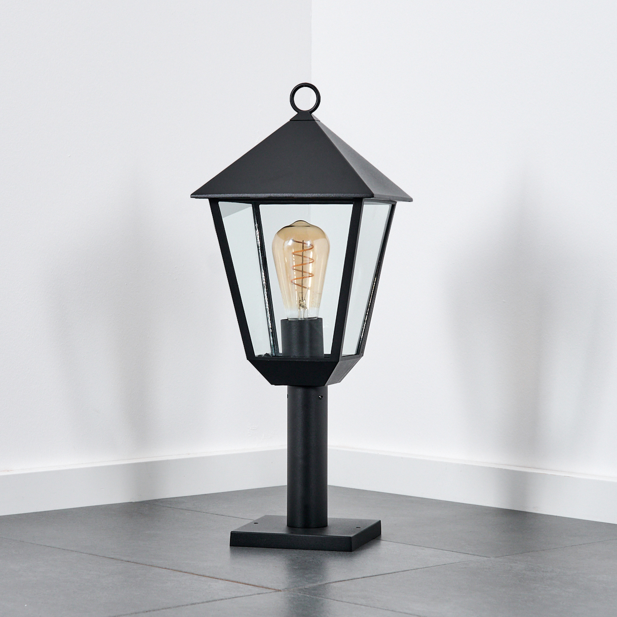 Schwarze Außenlampe mit klaren Glasscheiben und einer Glühbirne mit Wendel. Sie steht auf einem Fliesenboden in einer Ecke eines weißen Raumes.
