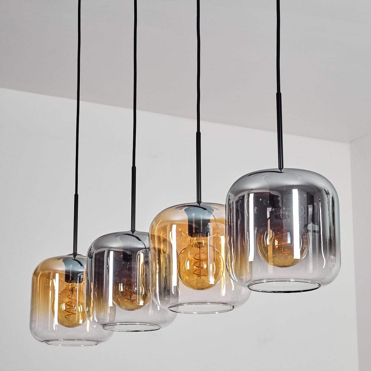 Vier Pendelleuchten mit Glasschirmen in verschiedenen Farben, jede mit einer sichtbaren Glühbirne. Die Decke ist weiß.