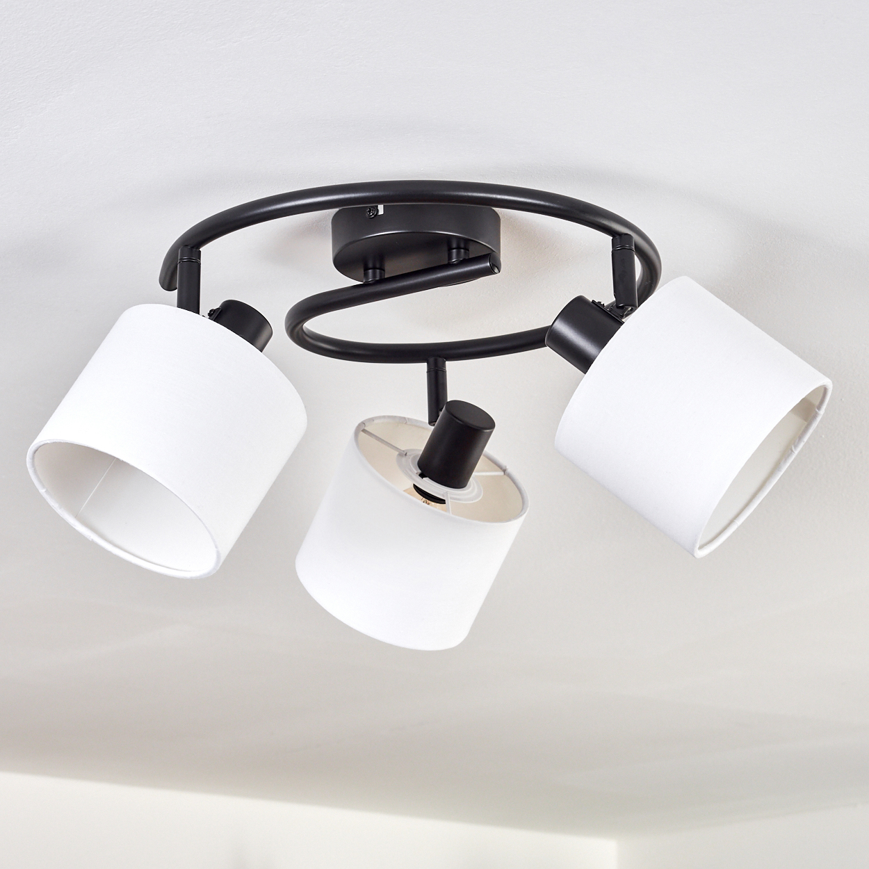 Eine Deckenleuchte mit drei weißen Lampenschirmen und schwarzen Halterungen, an einer weißen Decke montiert. Die Lampen sind abgewinkelt.