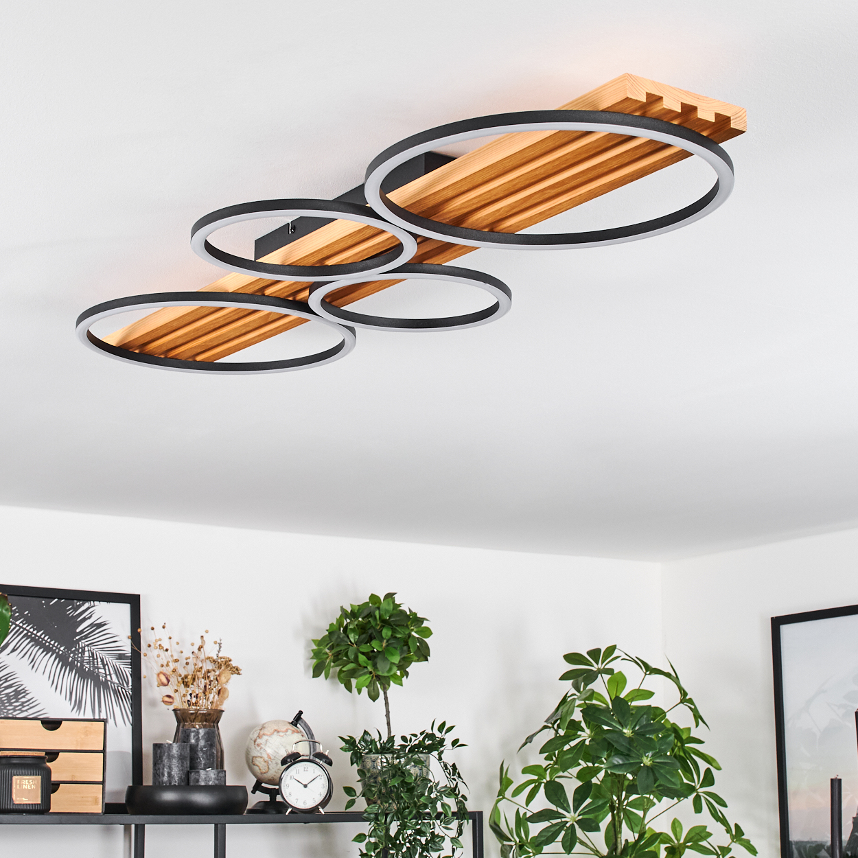 Moderne Deckenleuchte mit vier kreisförmigen Lichtern und Holzsockel. Darunter ein Regal mit Pflanzen und Deko-Artikeln.