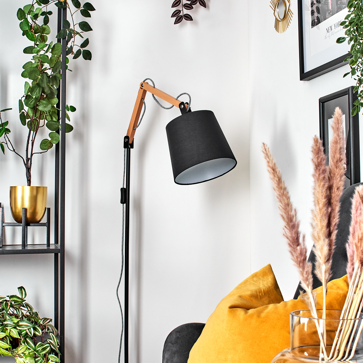 Eine schwarze Lampe mit Holzarm zeigt auf ein orangefarbenes Kissen. Pflanzen im Hintergrund mit schwarzem Regal.