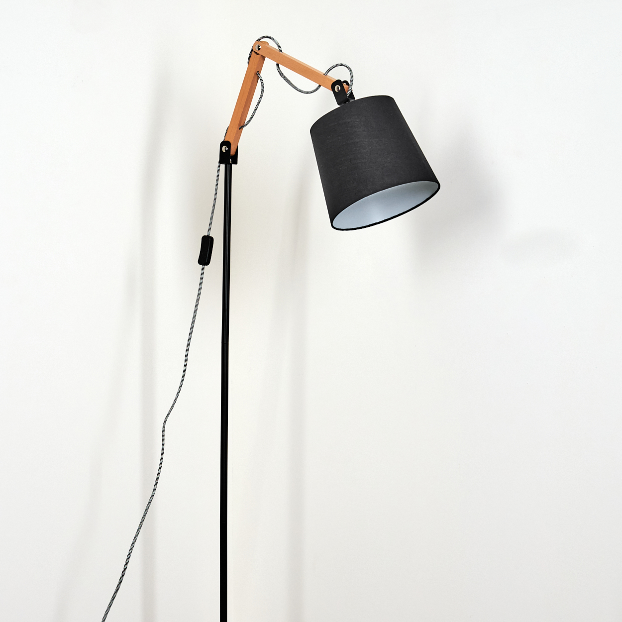 Eine schwarze und hölzerne Stehlampe steht vor einer weißen Wand. Sie hat einen schwarzen Schirm, einen Holzarm und ein schwarz-weißes Kabel.