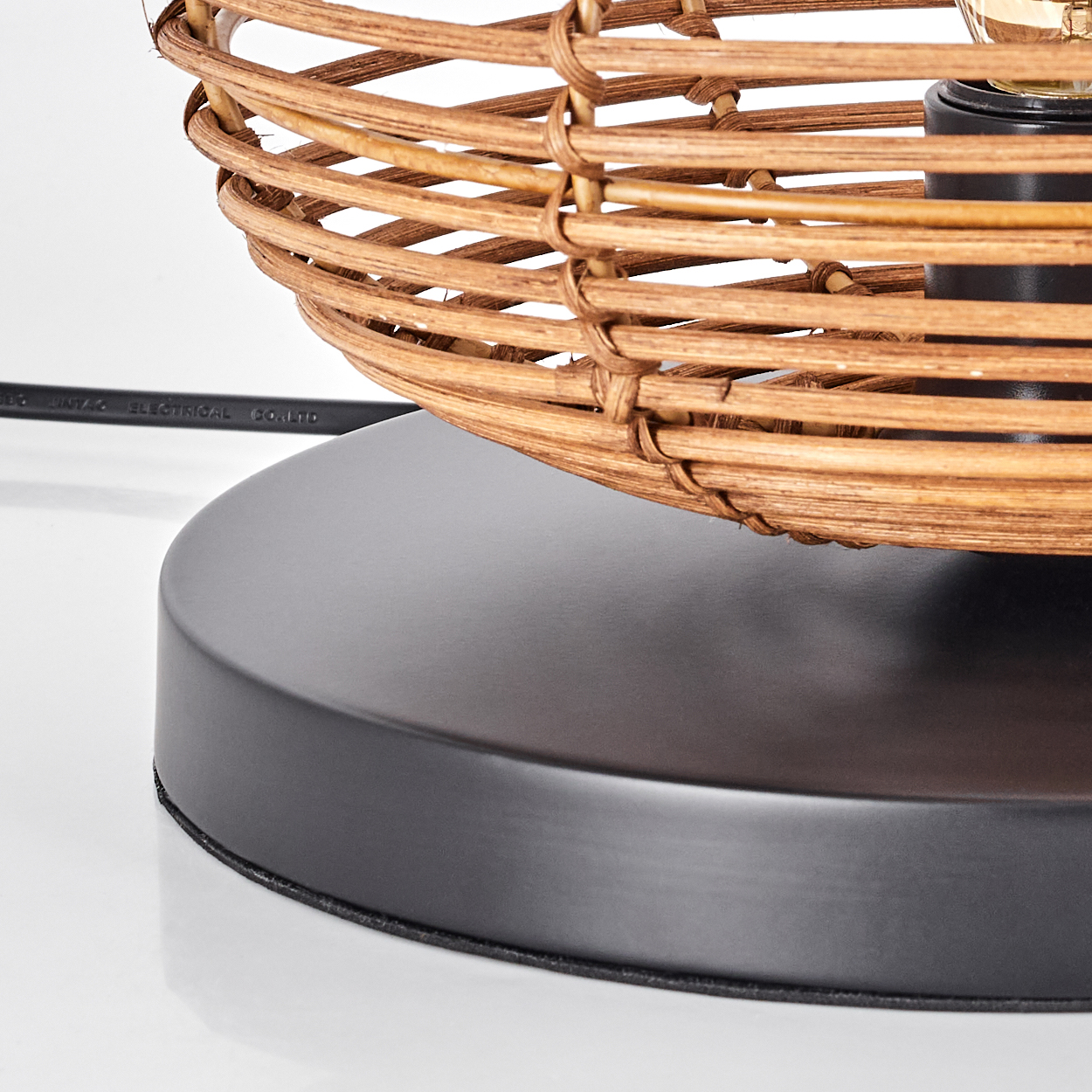 Nahaufnahme einer Lampe. Sie hat einen Lampenschirm aus geflochtenem Rattan, einen schwarzen Sockel und ein sichtbares Netzkabel. Der Hintergrund ist weiß.