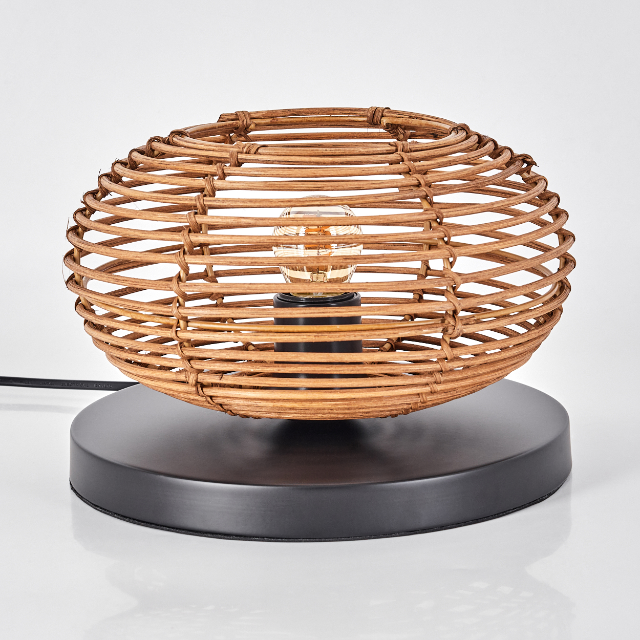Eine Lampe aus geflochtenem Rattan mit einer Glühbirne. Sie hat einen schwarzen Sockel und ein schwarzes Kabel. Der Hintergrund ist weiß.