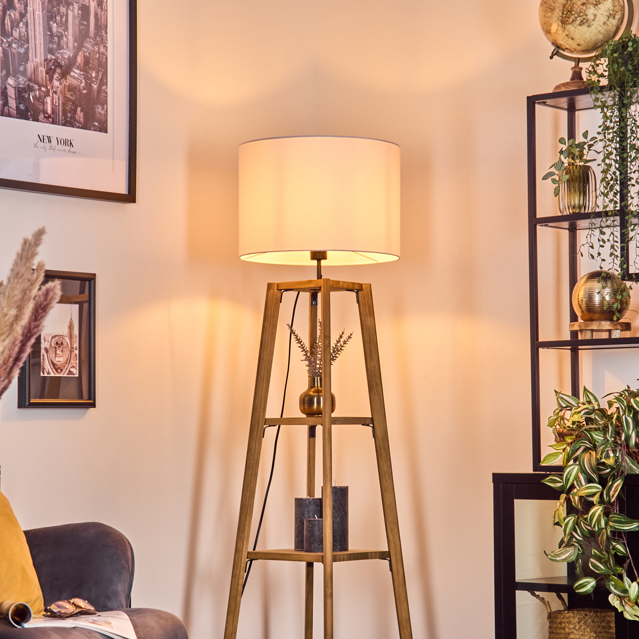 Eine Stehlampe mit beigem Lampenschirm steht in einem Raum. Im Hintergrund ein Regal mit Pflanzen und einem Globus. Ein New York-Poster an der Wand.