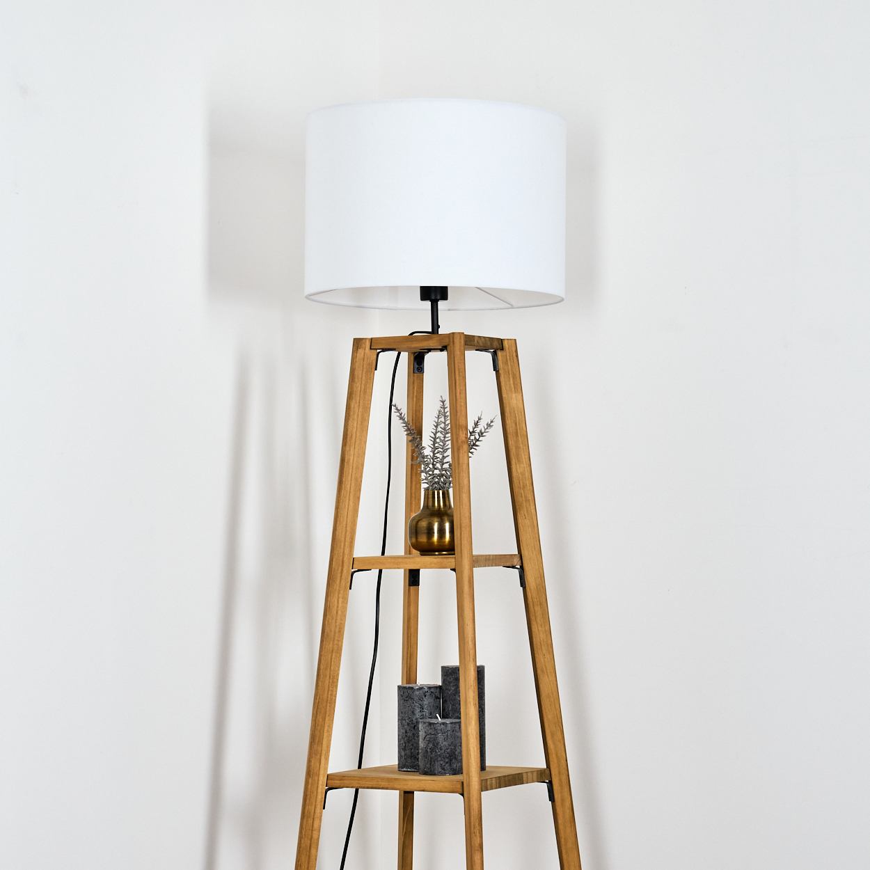 Eine Holzlampe mit weißem Lampenschirm. Sie hat eine kleine Vase mit Blumen und Kerzen auf ihren Regalen. Der Hintergrund ist weiß.