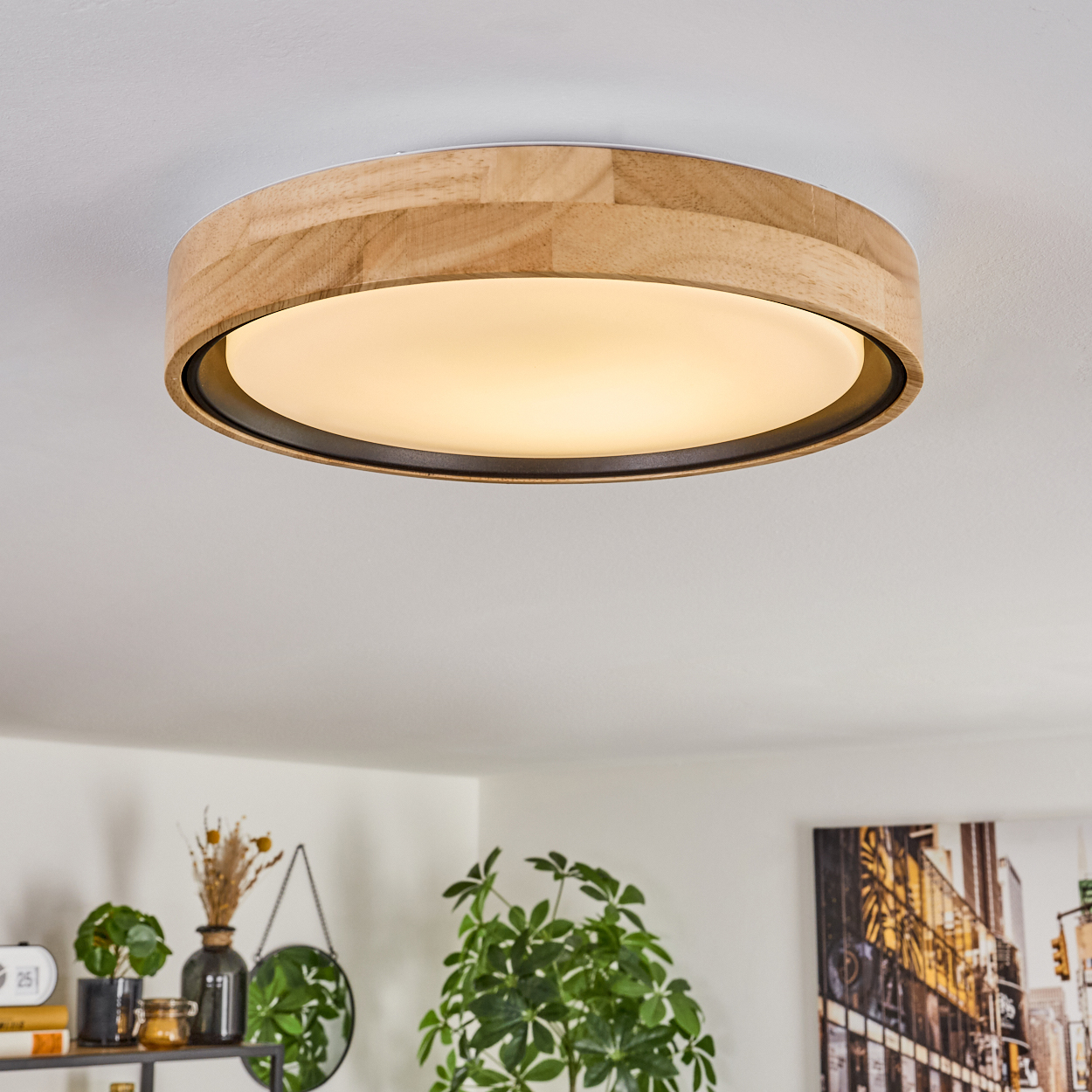 Eine Deckenleuchte aus Holz mit warmem Schein. Der Raum hat eine weiße Decke, ein Regal mit Pflanzen und ein Stadtbildgemälde.