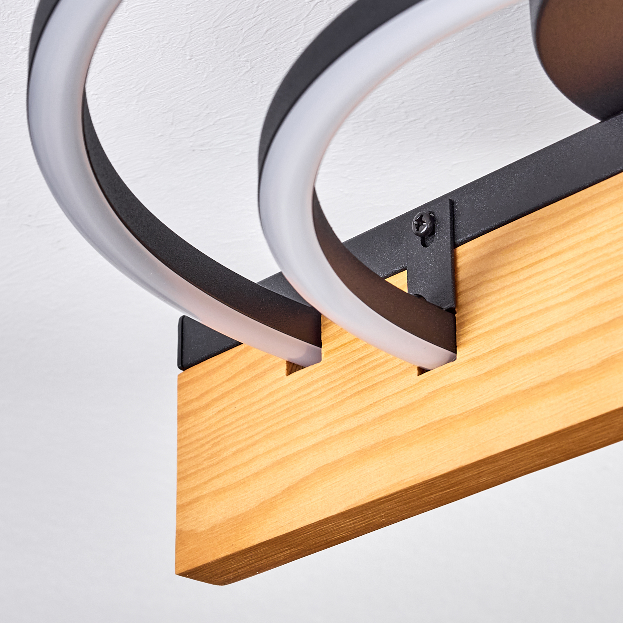 Nahaufnahme einer modernen Deckenleuchte. Sie zeigt einen Holzbalken und zwei gebogene, leuchtende weiße und schwarze Ringe an einer weißen Decke.