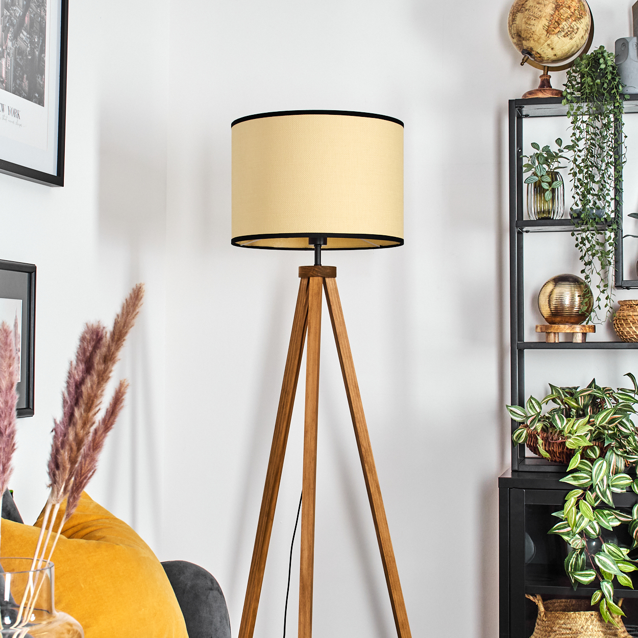 Eine Stehlampe mit beigefarbenem Lampenschirm und Holz-Dreibein steht in einem Raum mit Dekoration, darunter Pflanzen und ein Globus.