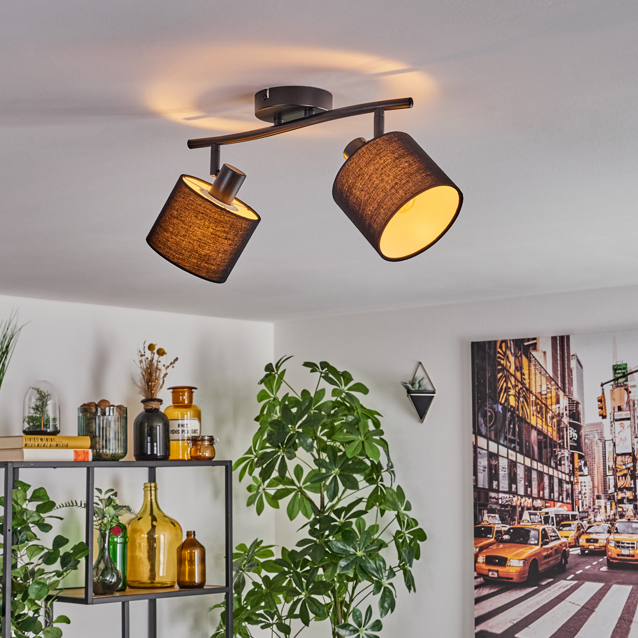 Zwei schwarze Lampen an einer Decke, die nach unten leuchten. Ein Regal mit Vasen und Pflanzen sowie ein New York City-Bild sind sichtbar.